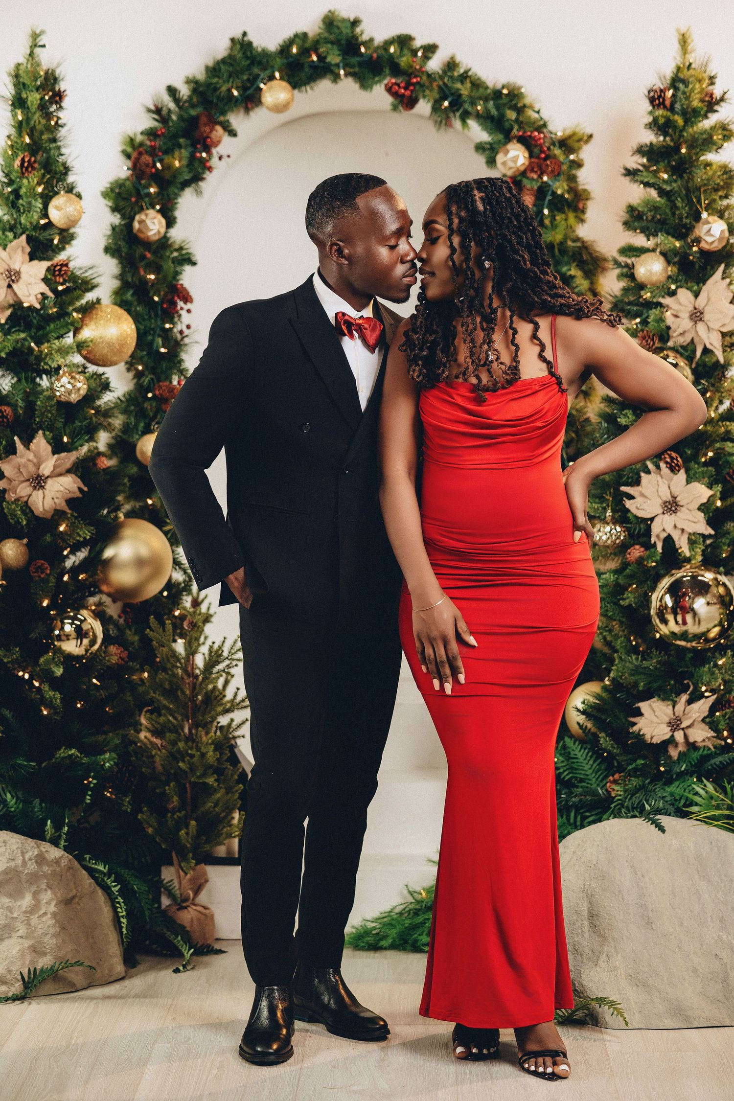 Couple sharing a kiss in red gown and black tuxedo before decorated Christmas tree arch.