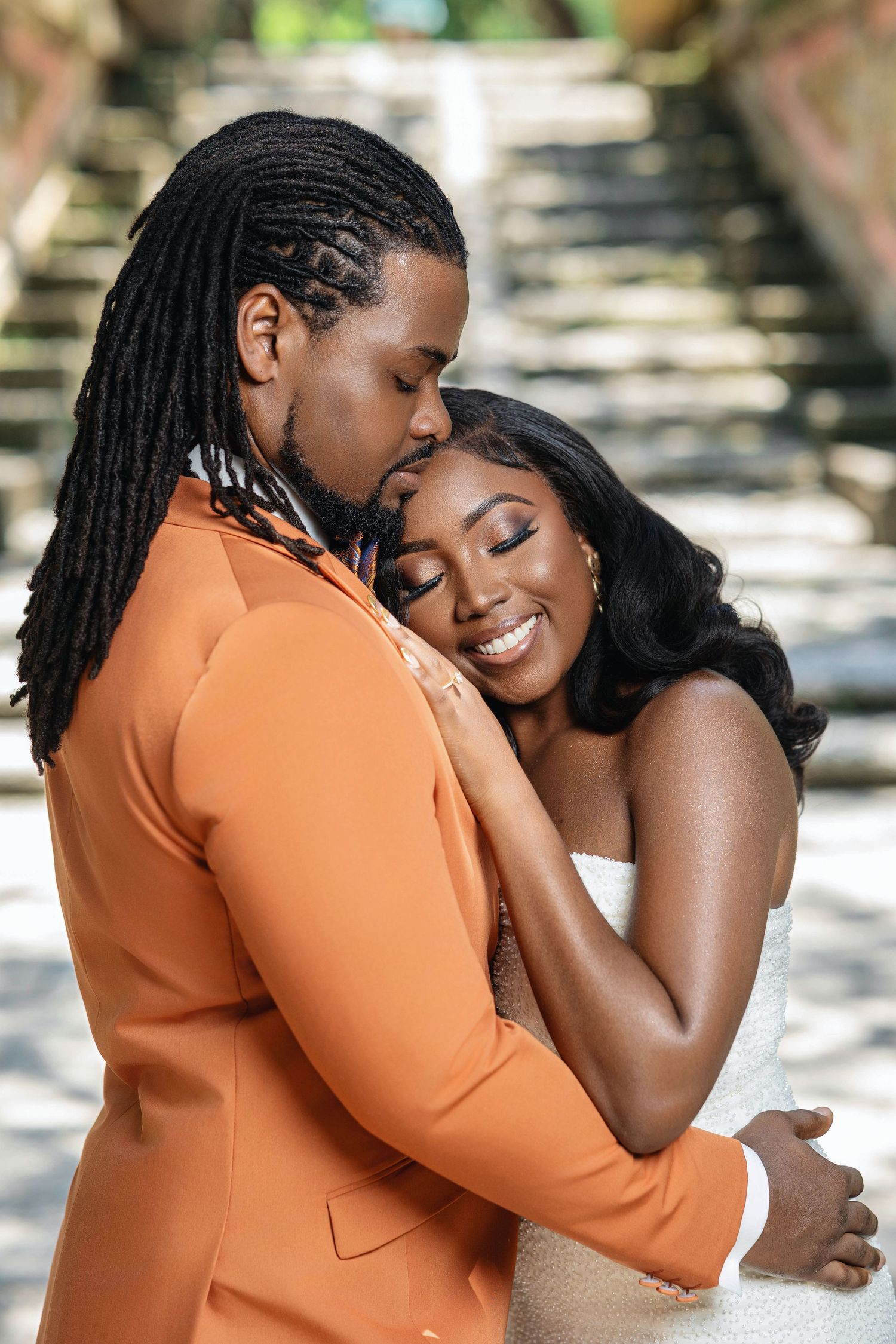 Groom in orange suit tenderly embraces smiling bride in white dress outdoors.