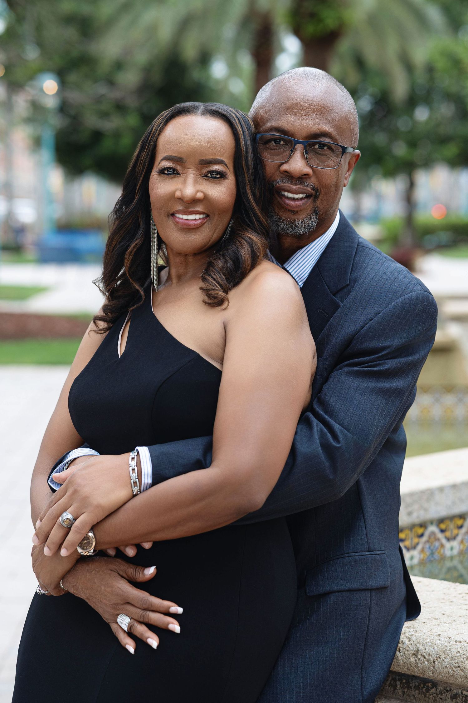 Elegant engaged couple embracing warmly outdoors, woman in black dress with diamond ring, man in navy suit.
