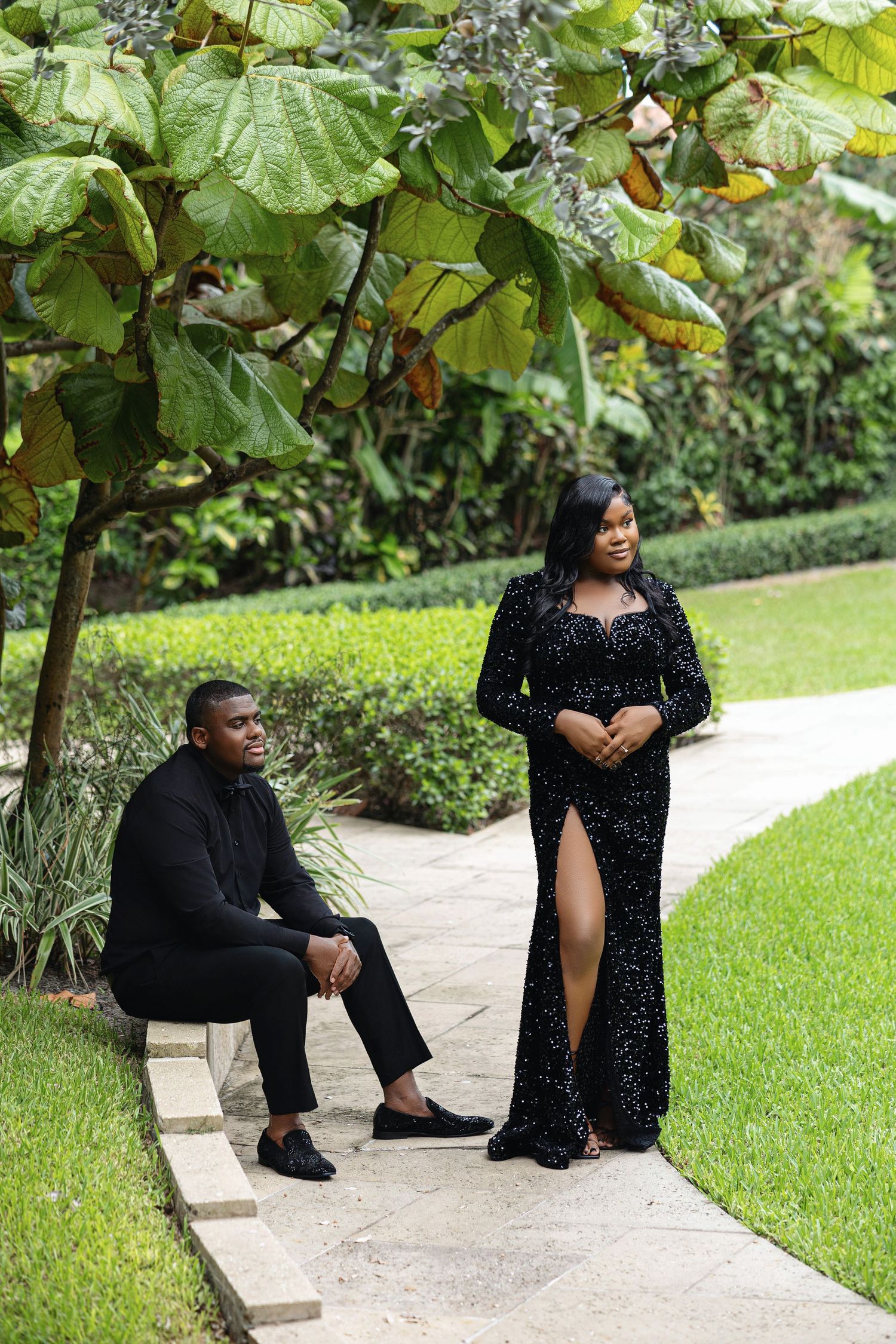 Couple dressed in black formal attire posing on a garden pathway surrounded by lush green tropical foliage.