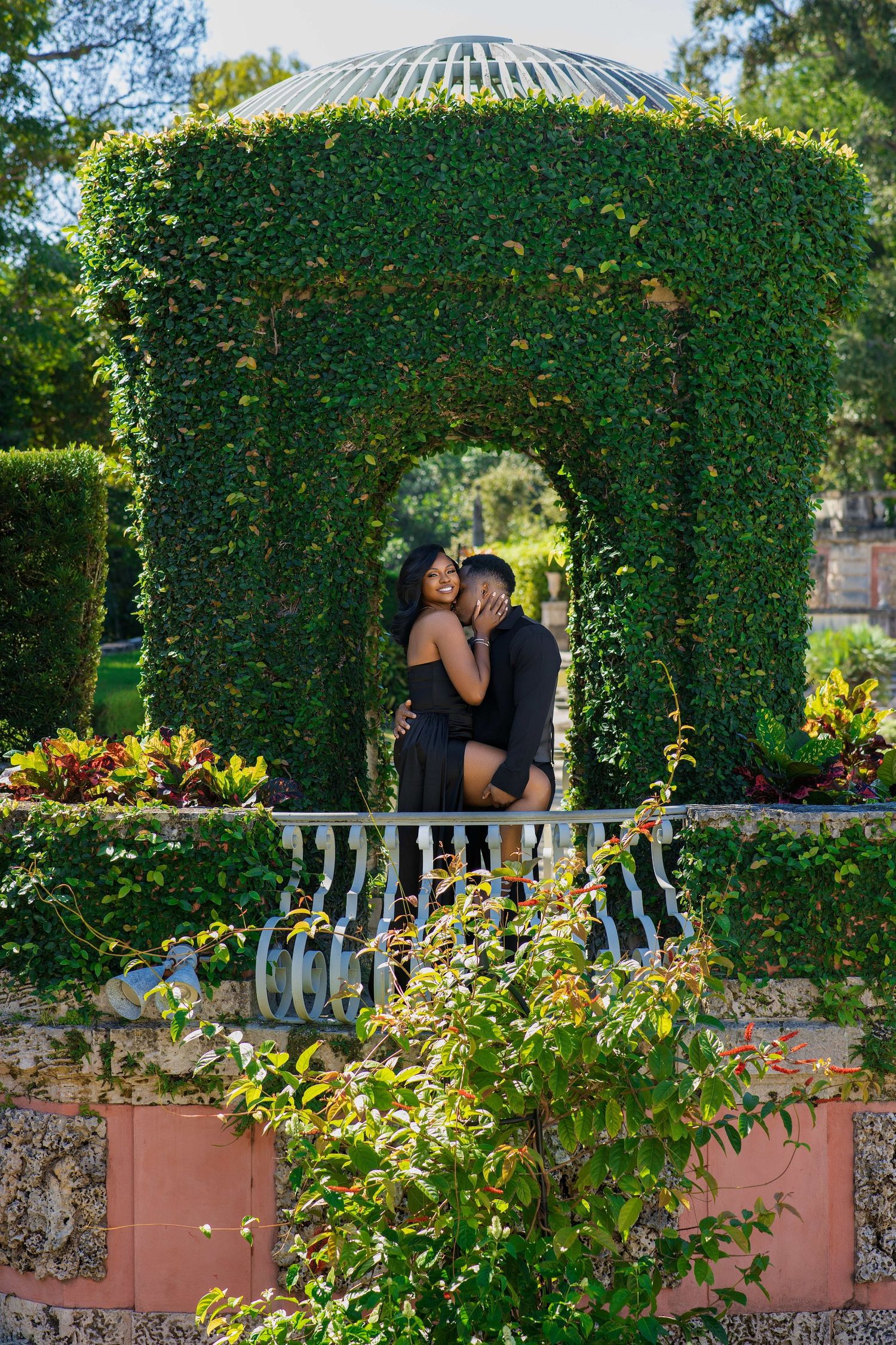 Romantic Black couple embracing inside ivy-covered gazebo arch surrounded by lush garden greenery.
