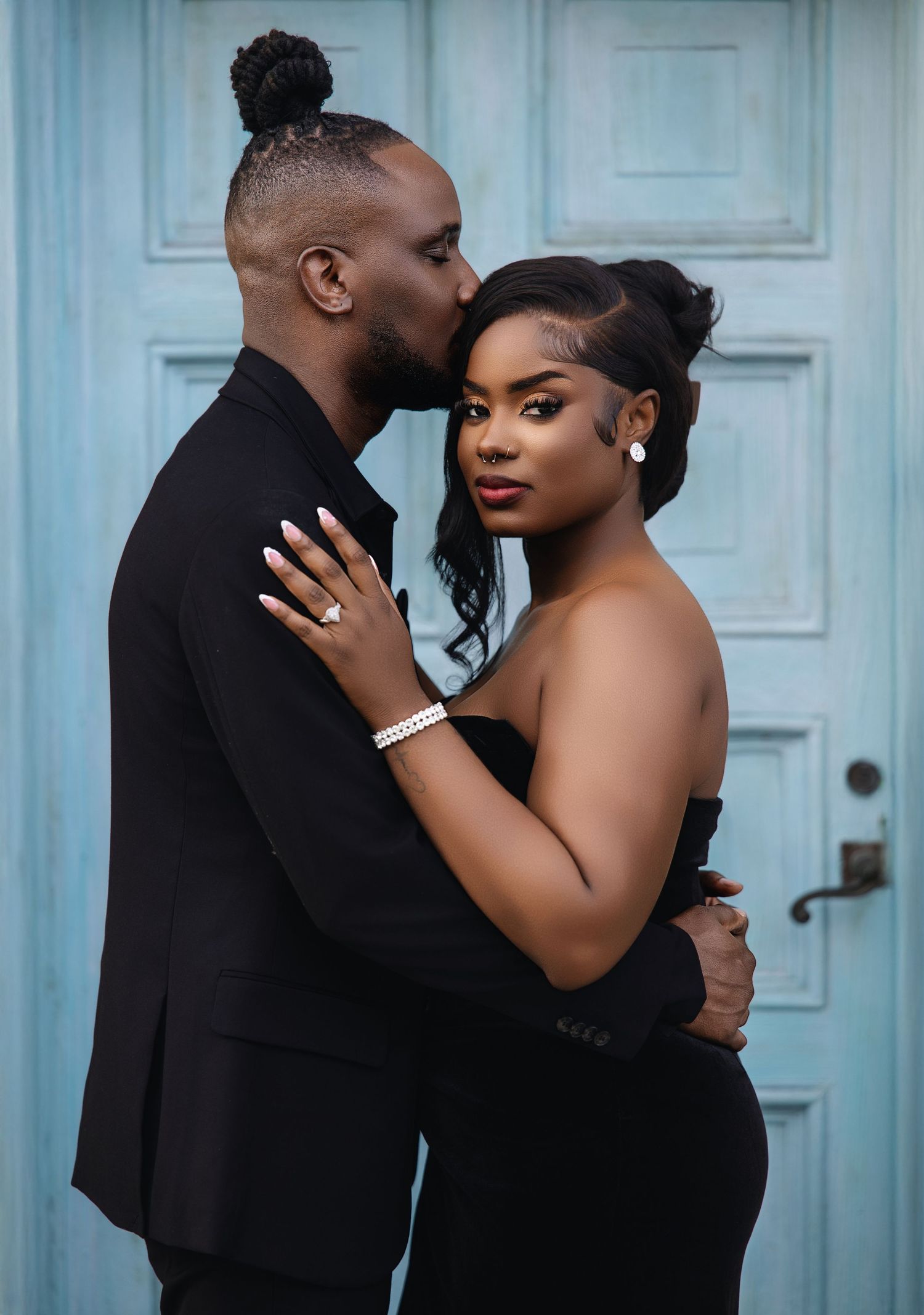 Man in black suit kissing woman in strapless black gown before a teal door.