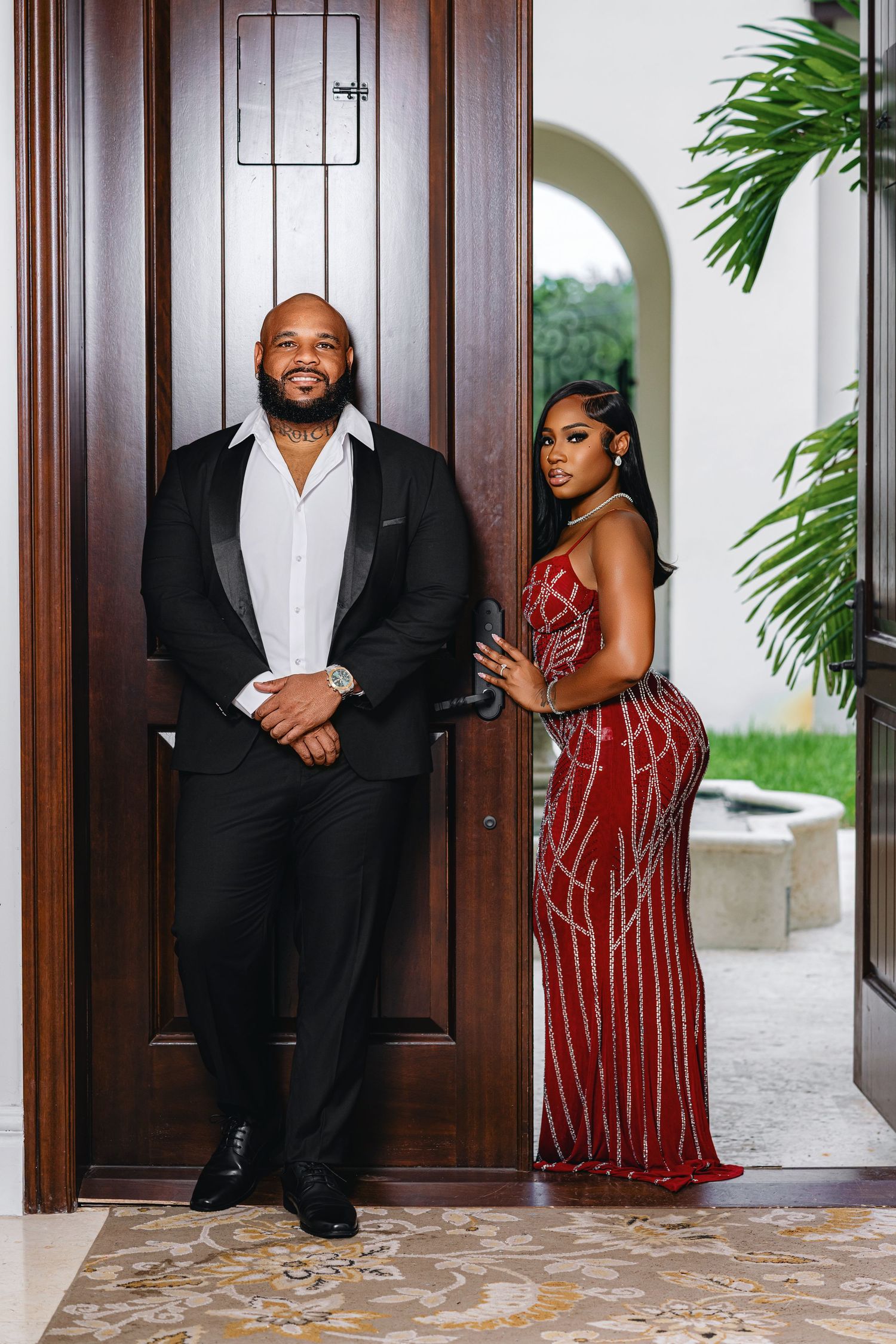 Couple poses at grand wooden entrance, man in black tuxedo and woman in red sequined form-fitting gown.
