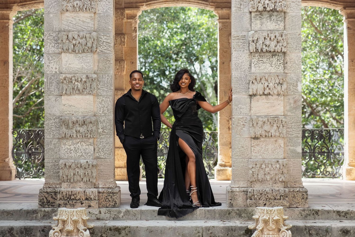 Stylish Black couple posing in all-black formal outfits between ornate marble columns at historic venue.