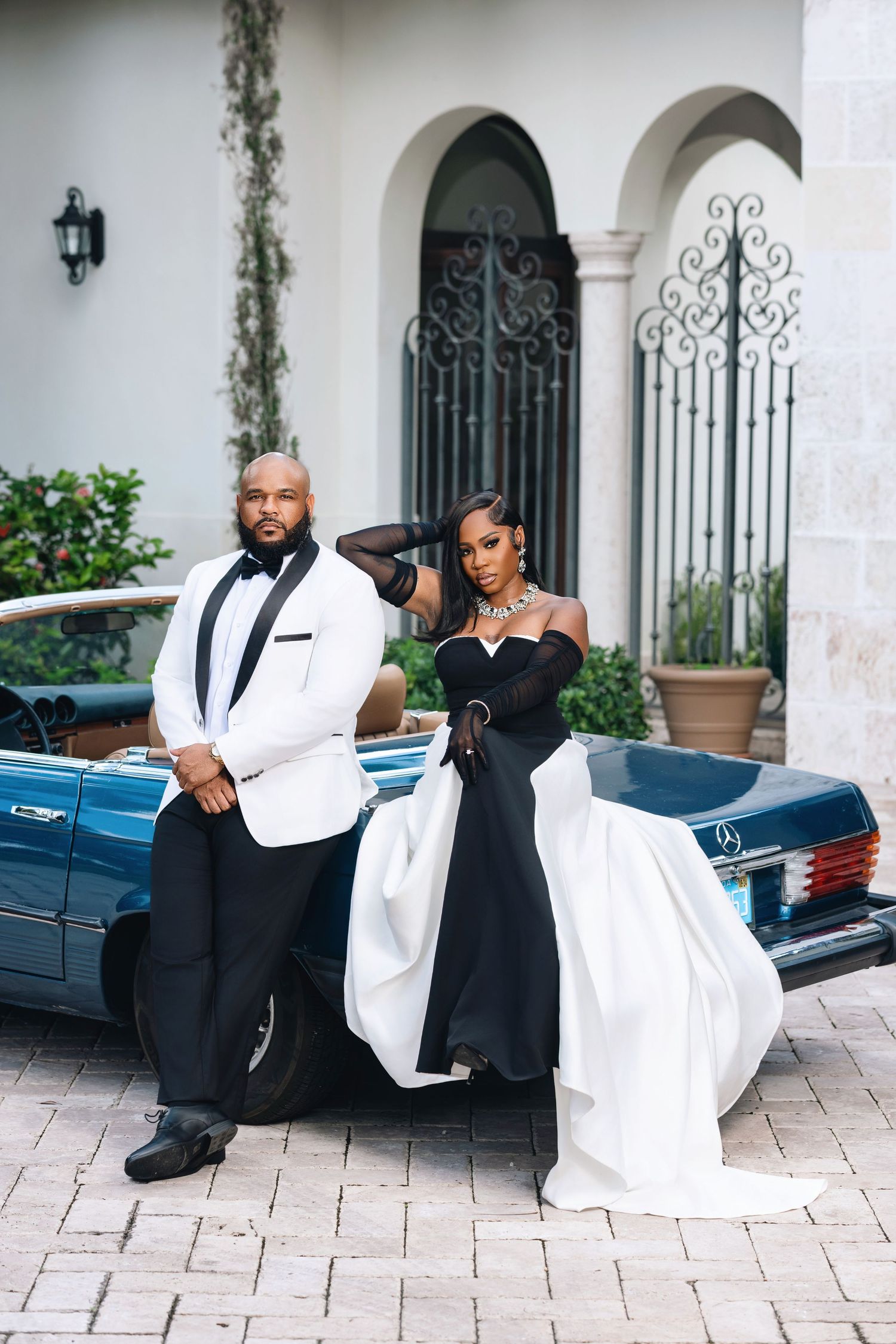 Glamorous couple in black and white formal wear pose beside a classic vintage blue Mercedes convertible.