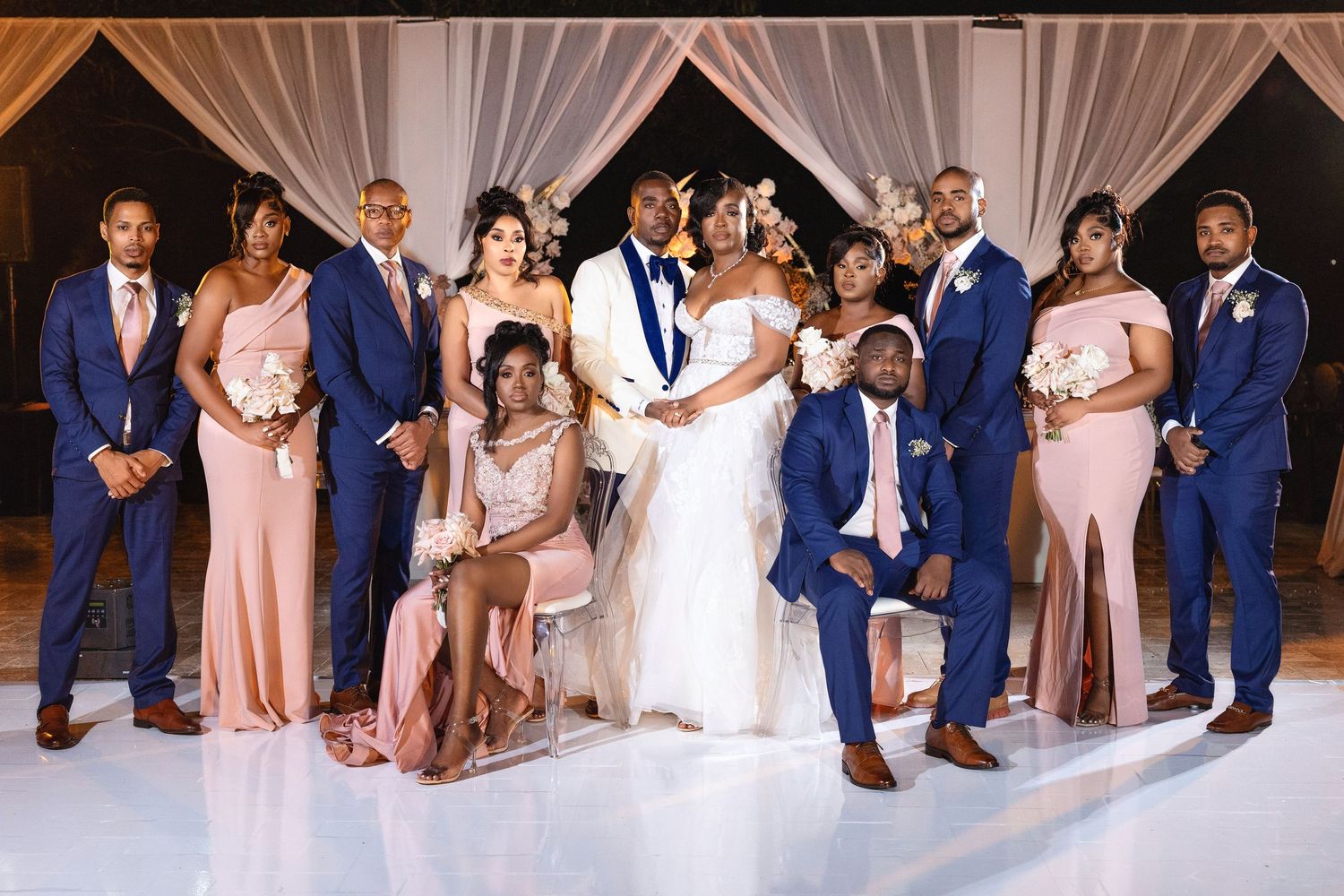 Wedding party poses together, bridesmaids in blush gowns and groomsmen in navy suits flanking the bride and groom.