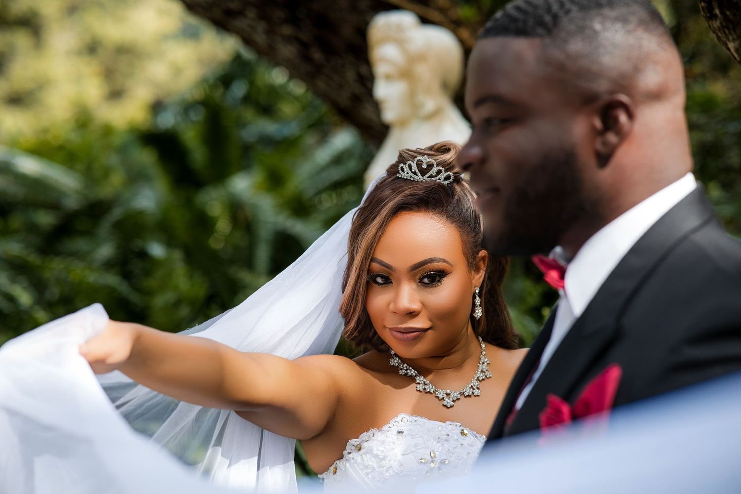 A bride in a white gown and tiara adjusts her veil outdoors, with a groom in a suit beside her.