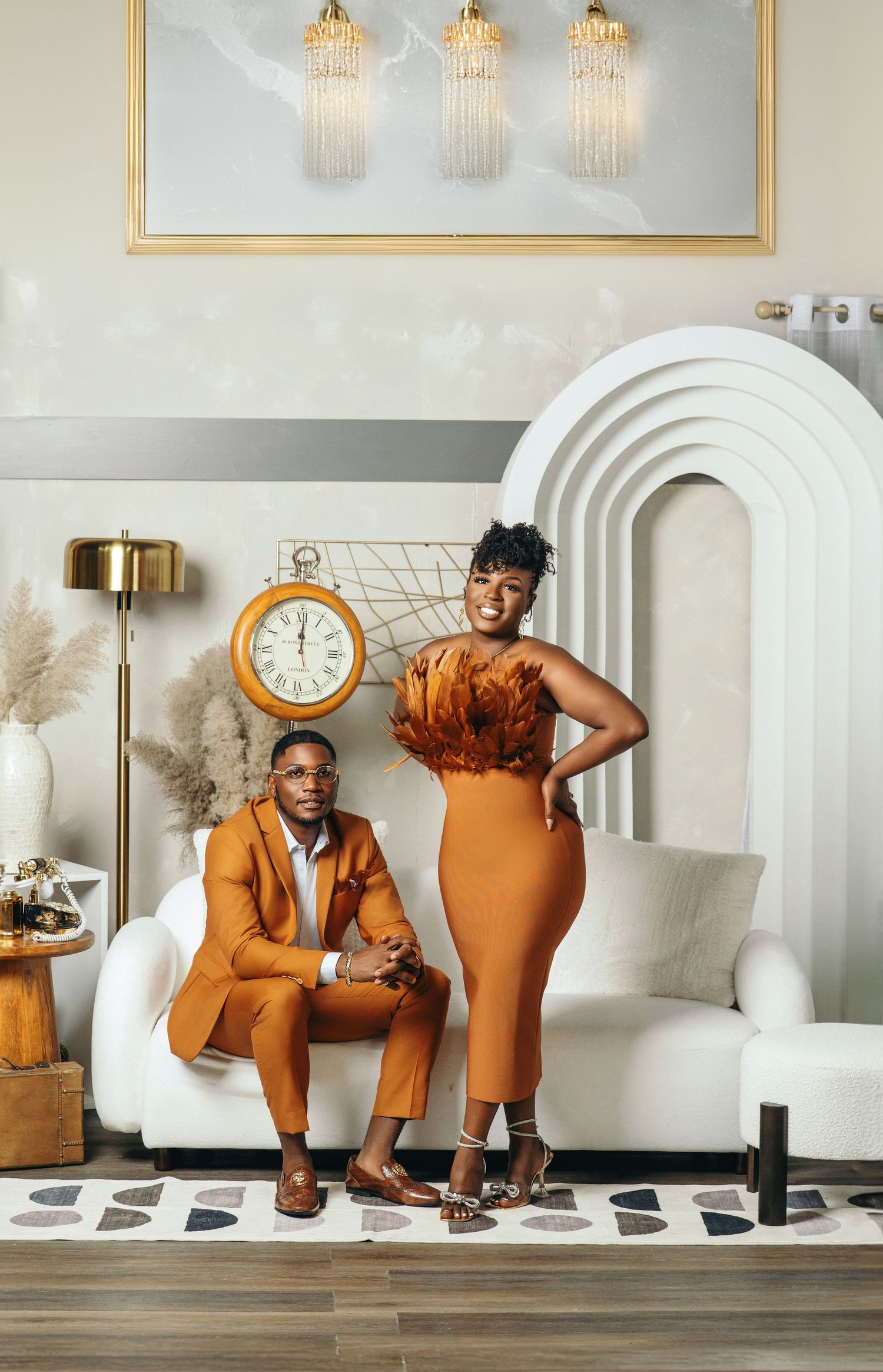 Couple posing in coordinating burnt orange outfits with feather top in a chic white interior.