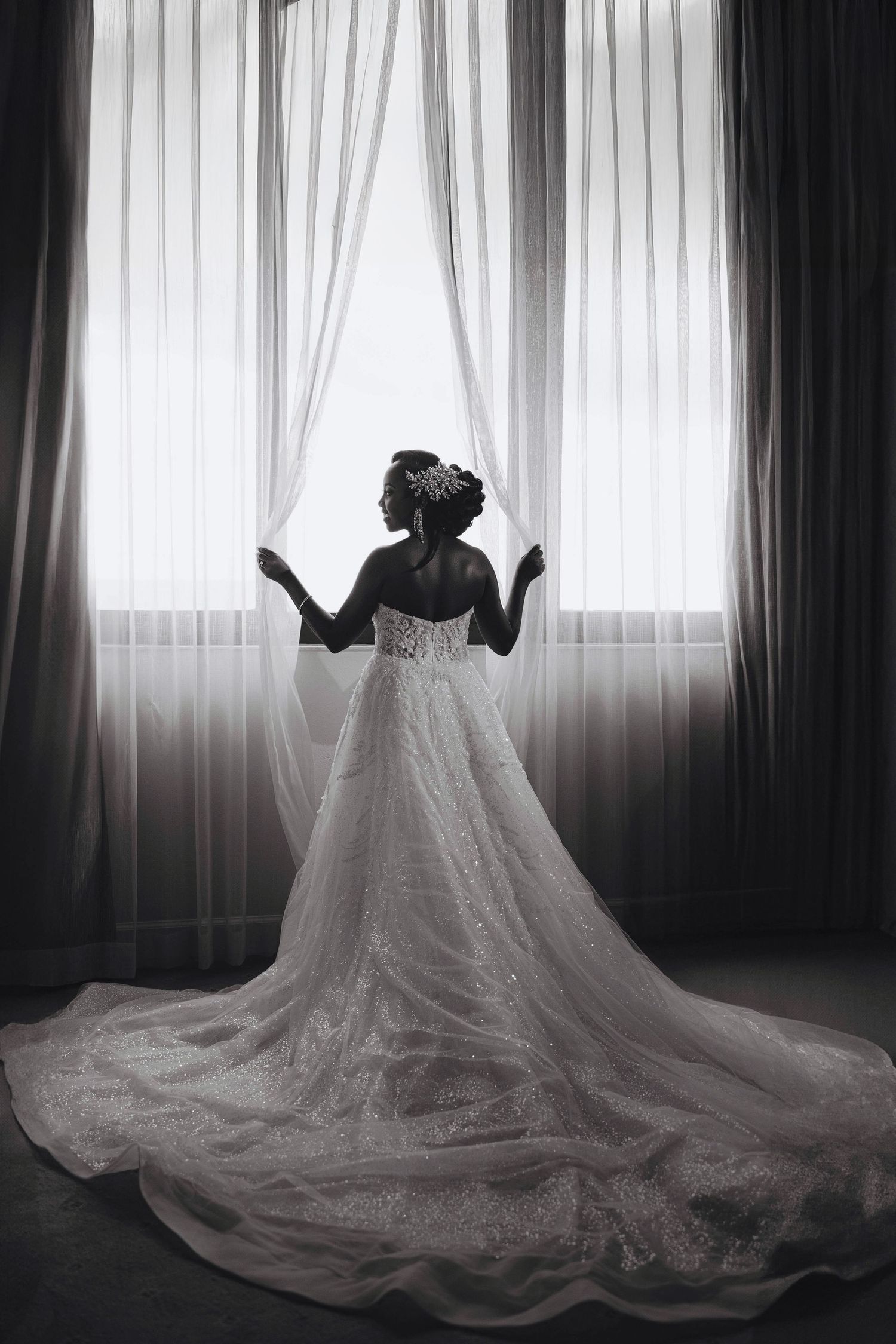 Black and white photo of a bride in a stunning ball gown with long train standing at a window.