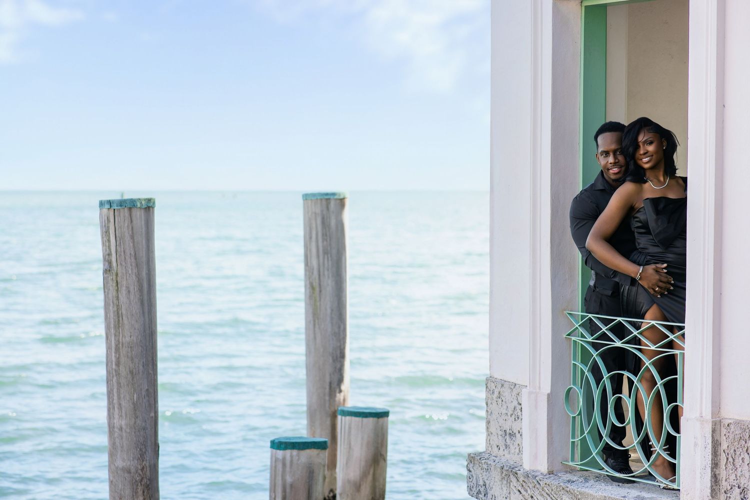 Couple in black outfits embracing in waterfront balcony doorway with ocean view.