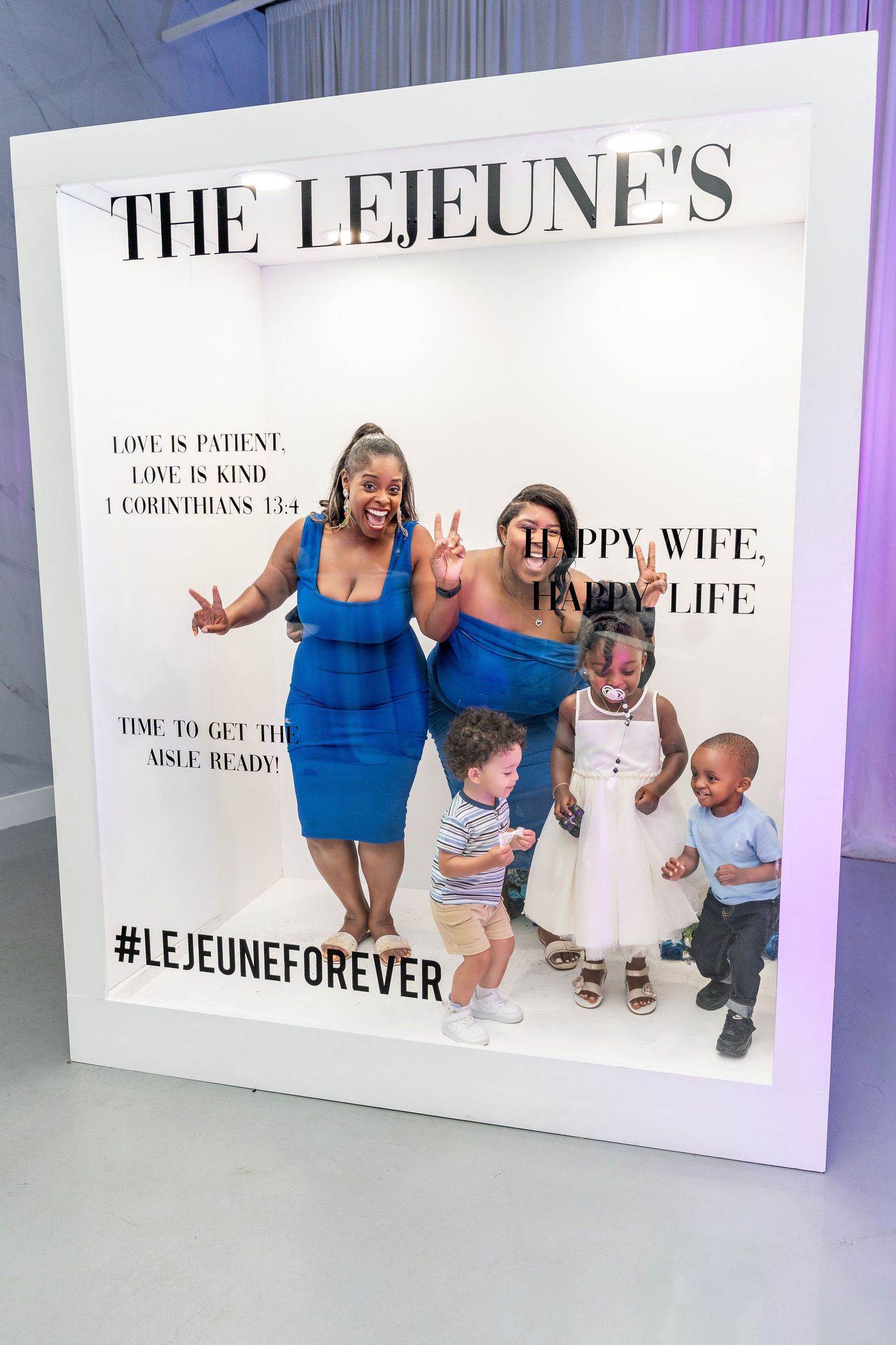 Two women and three young children pose inside The Lejeune's wedding photo booth with hashtag display.