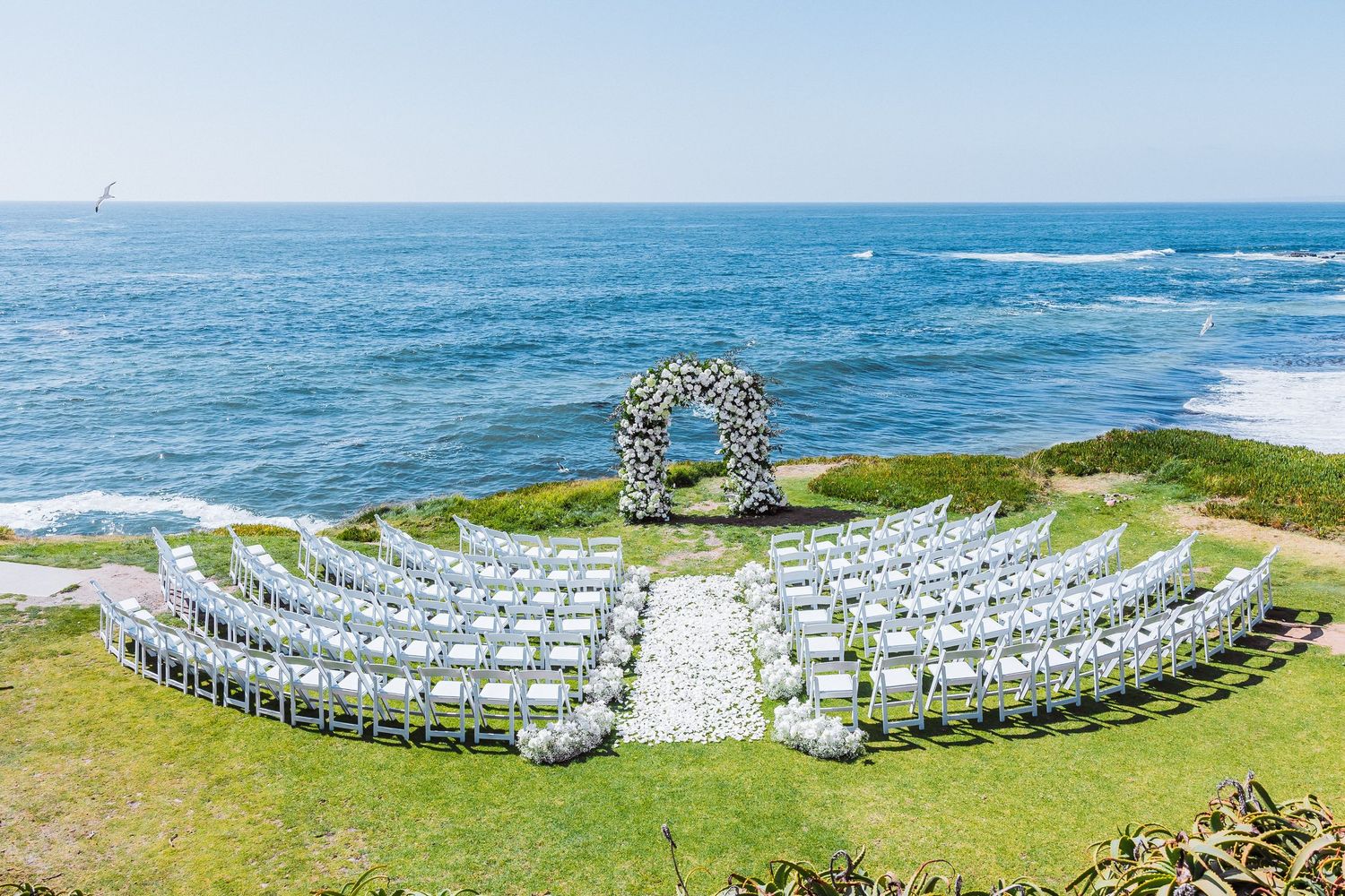 Outdoor oceanfront wedding ceremony setup with white chairs and floral arch overlooking the Pacific Ocean.