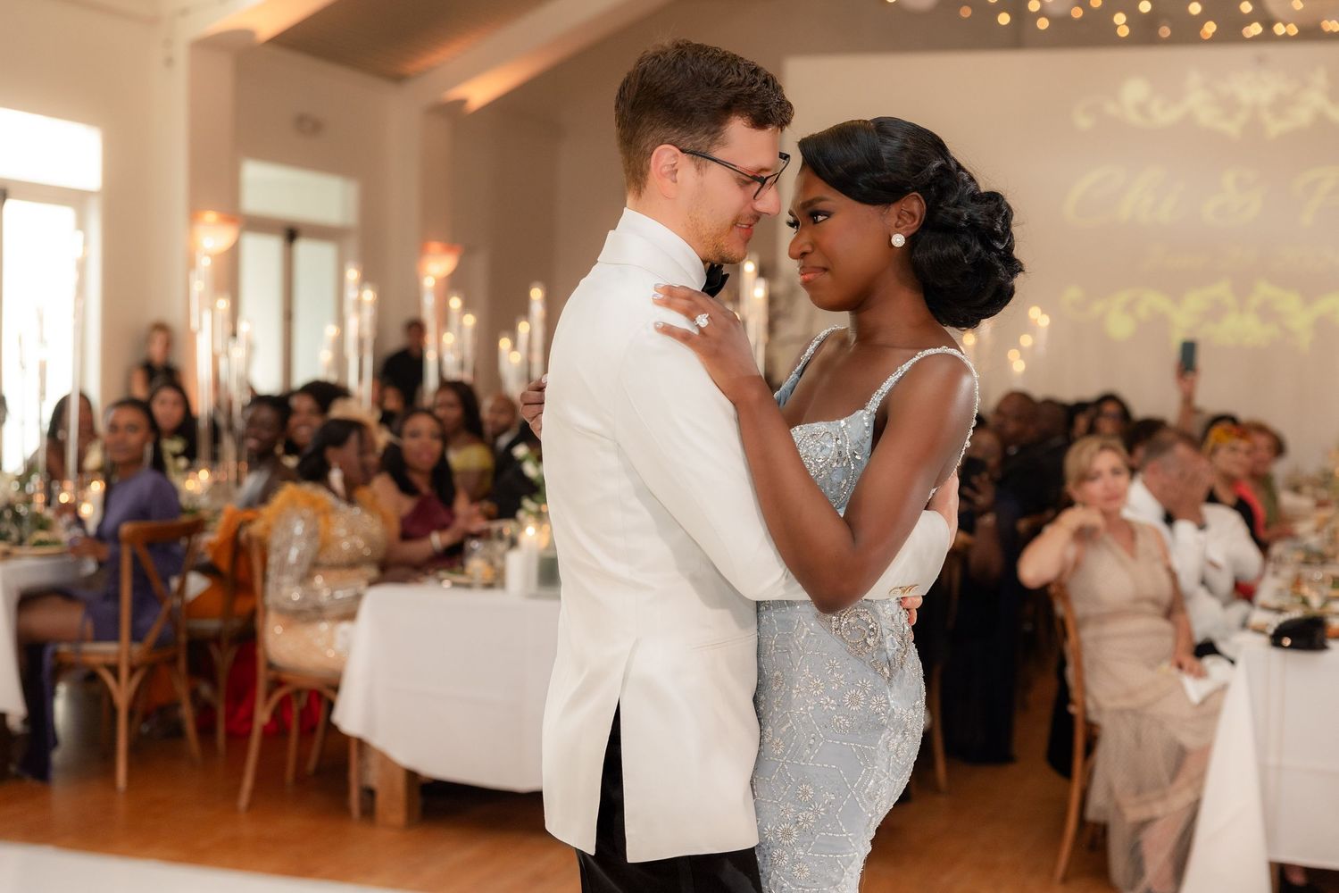 Couple sharing first dance at elegant wedding reception, groom in white tuxedo, bride in silver gown, guests seated in background.