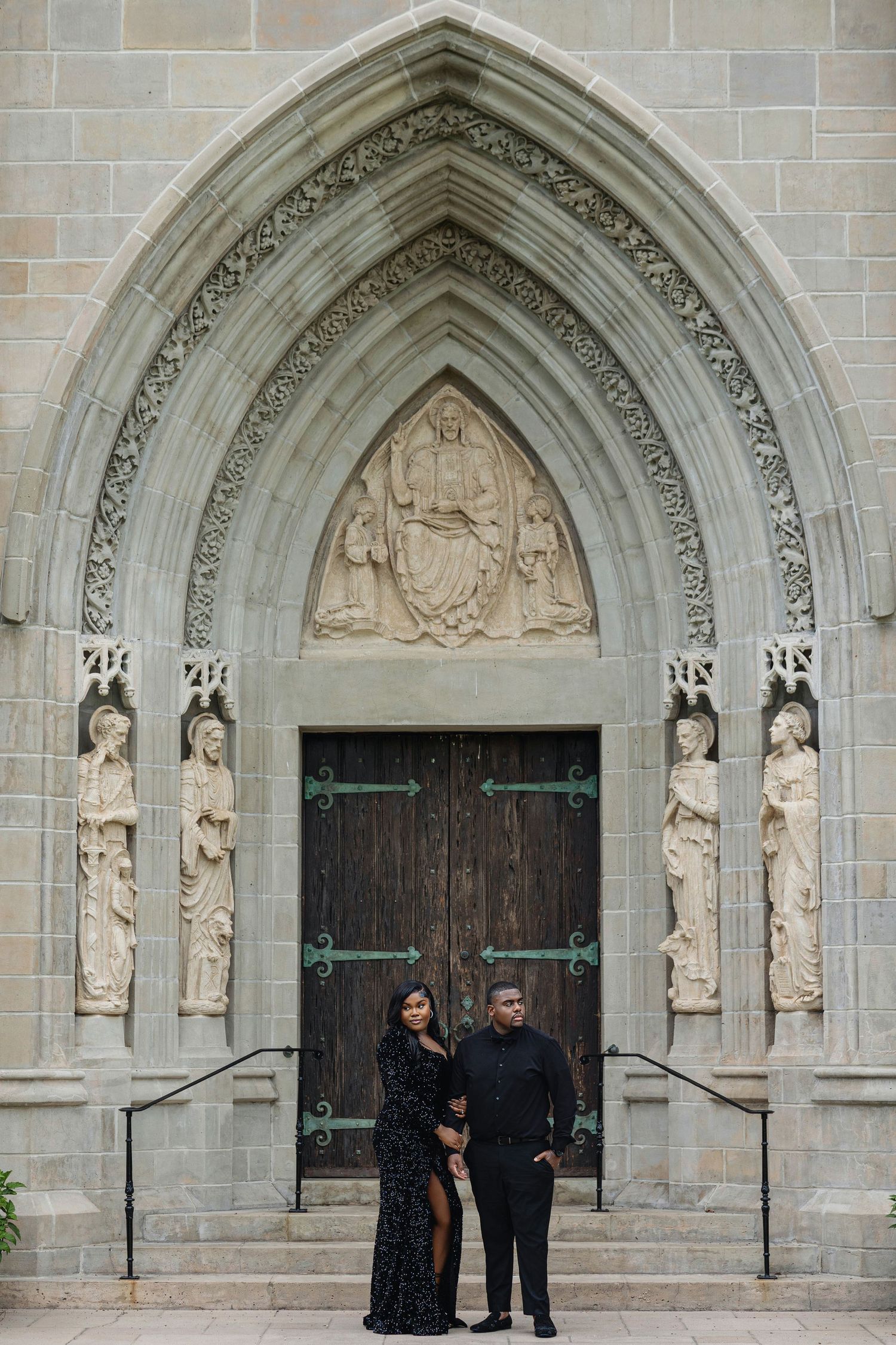 Couple in matching black outfits posing before ornate Gothic church entrance with stone carvings and wooden doors.