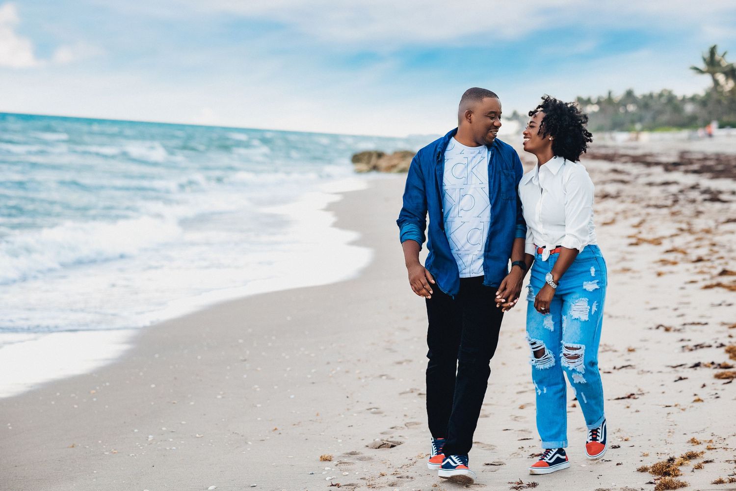 Happy couple walking hand in hand along a sandy beach with ocean waves in the background.