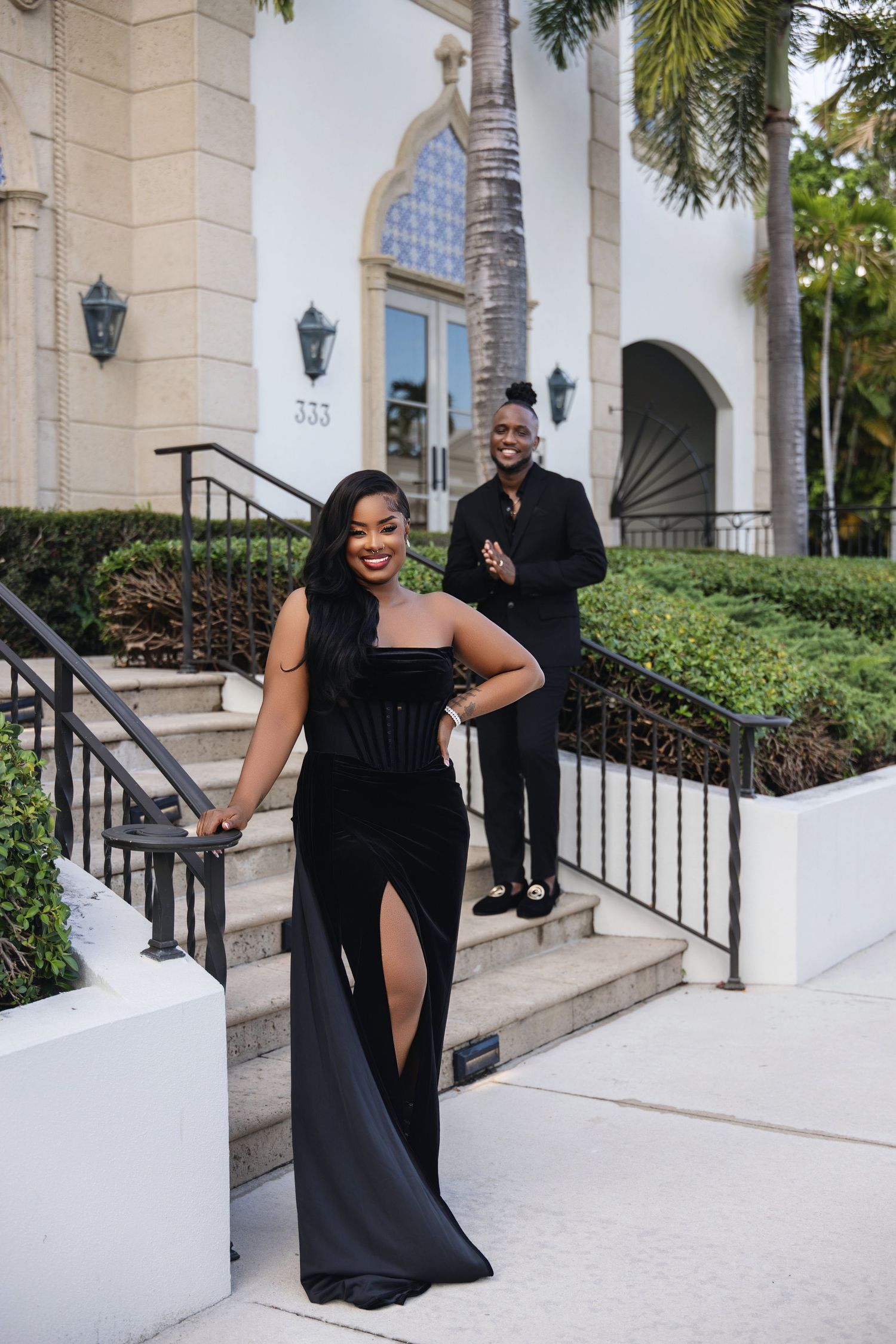 Woman in black gown poses on steps while man in black suit stands behind.