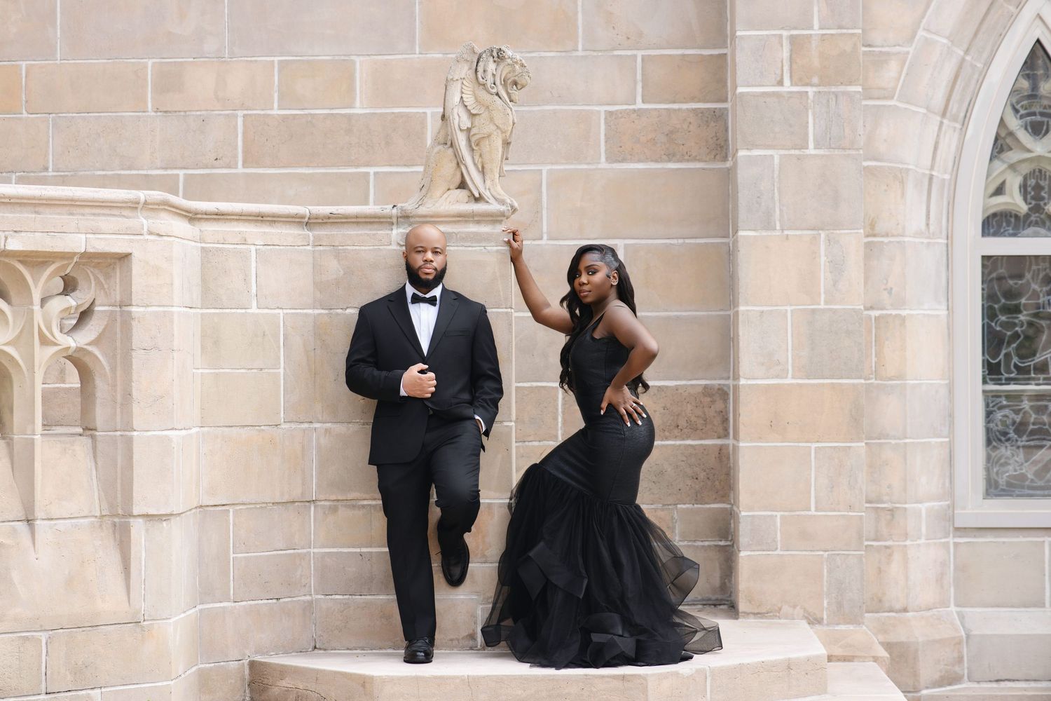 Stylish couple in black formal wear posing beside stone lion sculpture on castle-like stone steps.
