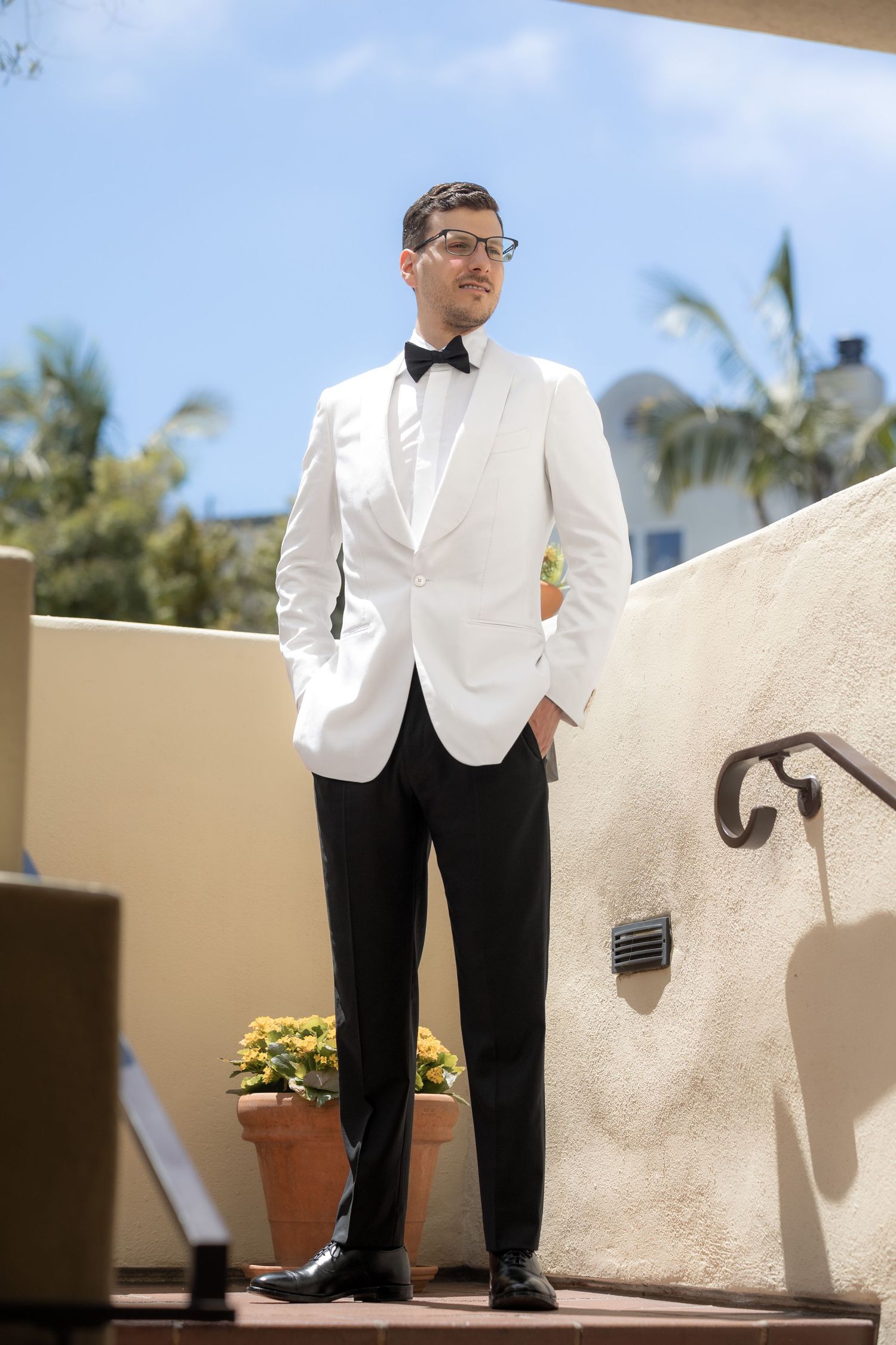 A man in a white tuxedo jacket, black bow tie, and trousers stands confidently on an outdoor terrace.
