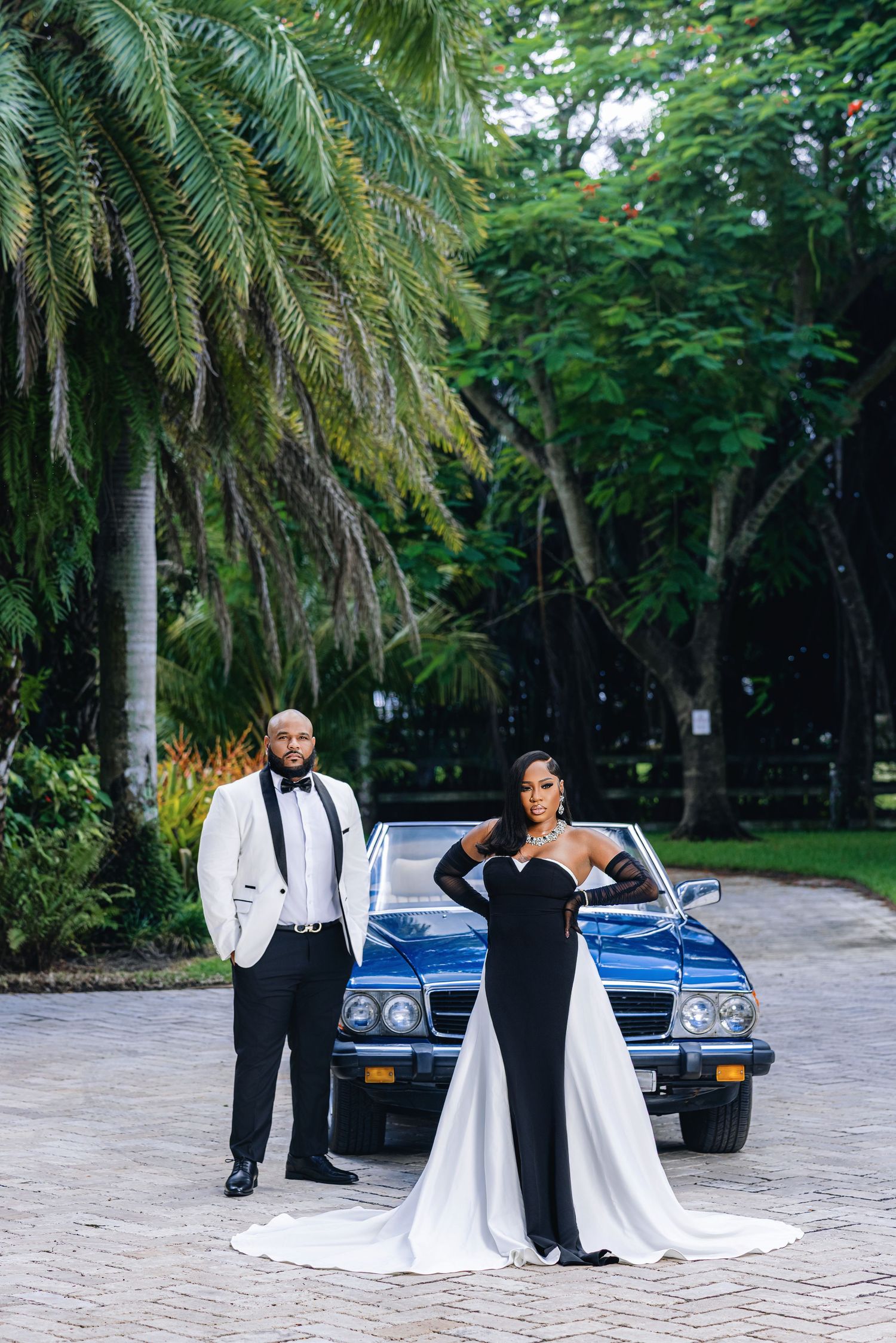 Glamorous wedding couple posing before vintage blue convertible surrounded by lush tropical palm trees.