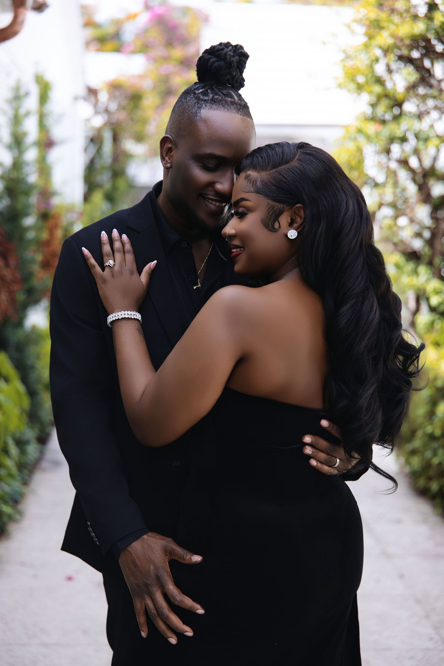 A stylish Black couple embraces intimately in a lush garden, both wearing coordinating black outfits.