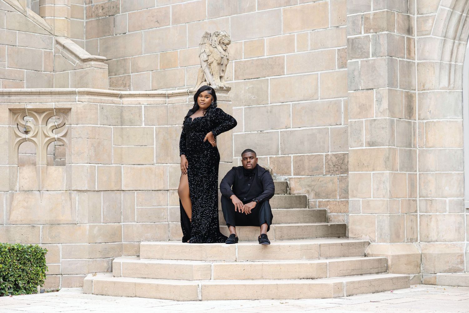 Elegant couple in black attire posing on stone steps of a Gothic architecture building exterior.
