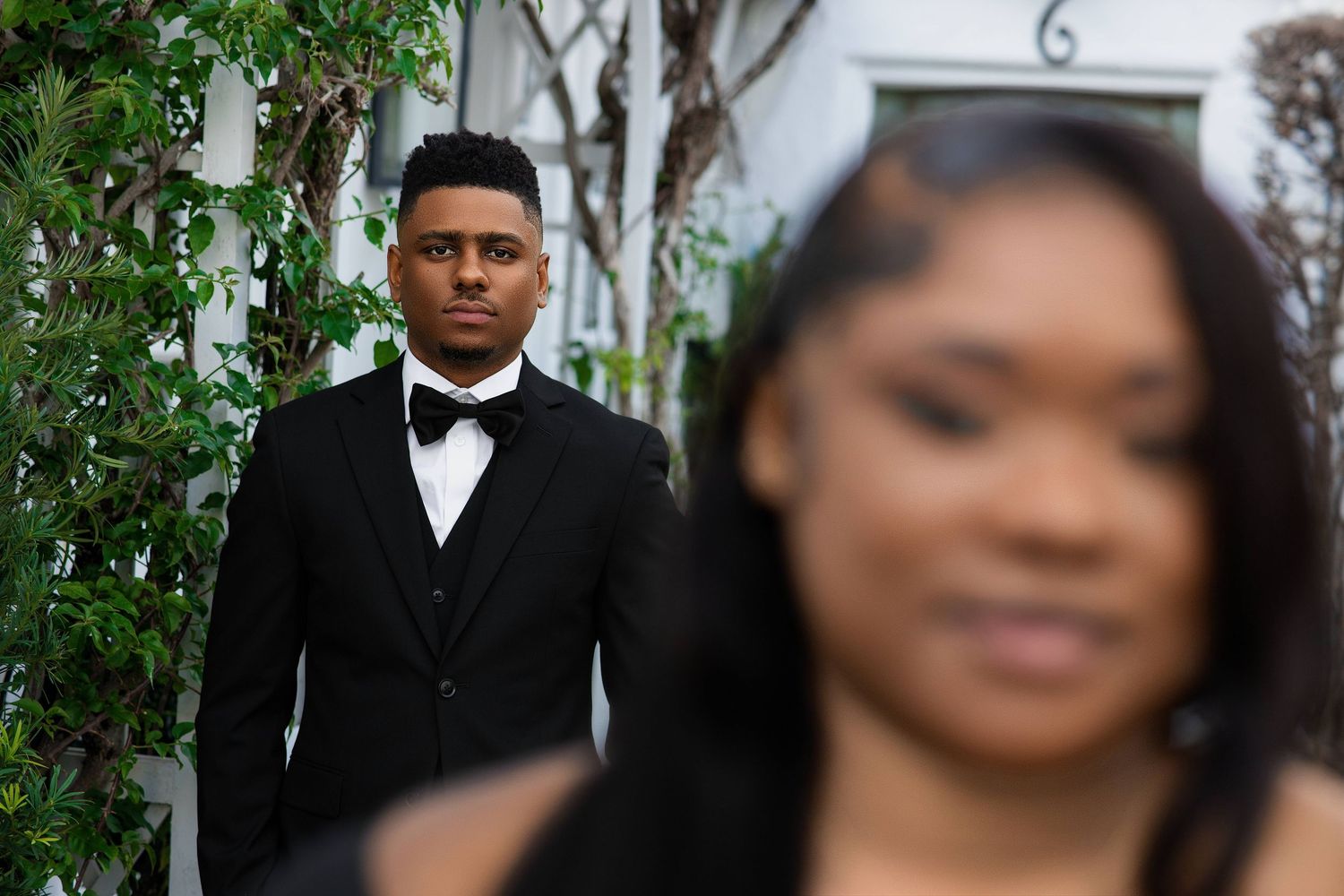 Man in black tuxedo standing in garden with woman in foreground, shallow focus.