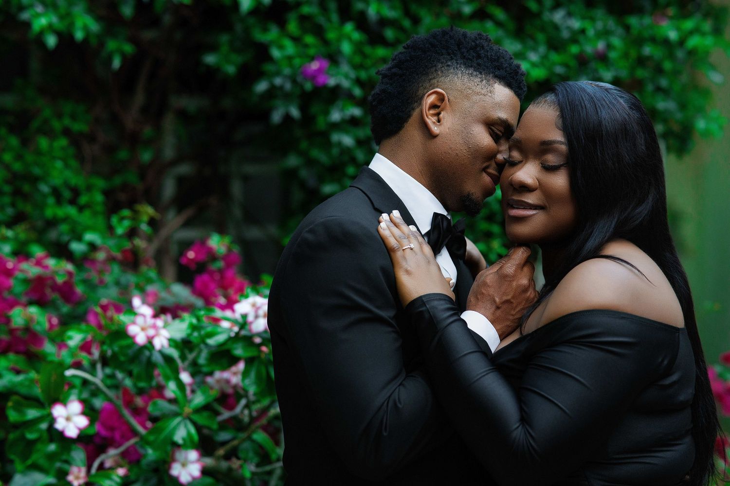 A couple in black formal wear share an intimate, romantic moment surrounded by vibrant garden flowers.