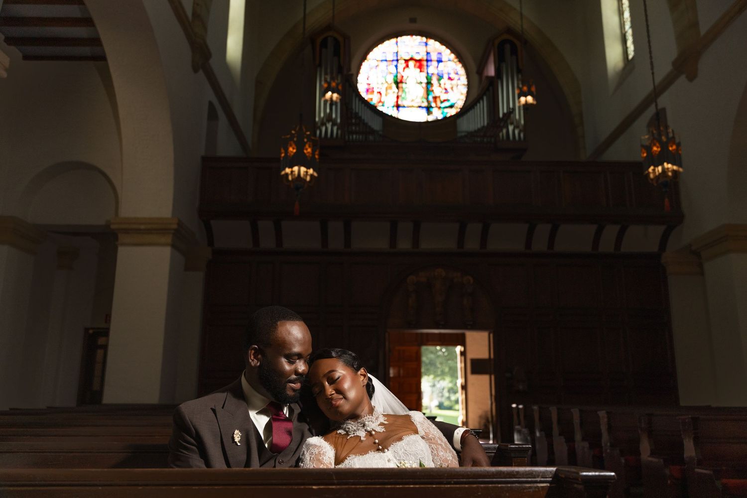 Bride and groom sitting together in a church pew, with stained glass window and organ pipes in the background.