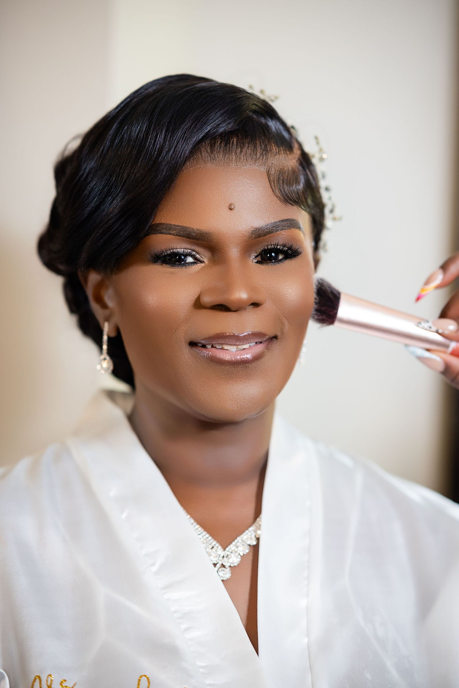 A bride getting makeup applied with a brush while wearing a white robe, diamond jewelry, and elegant updo hairstyle.