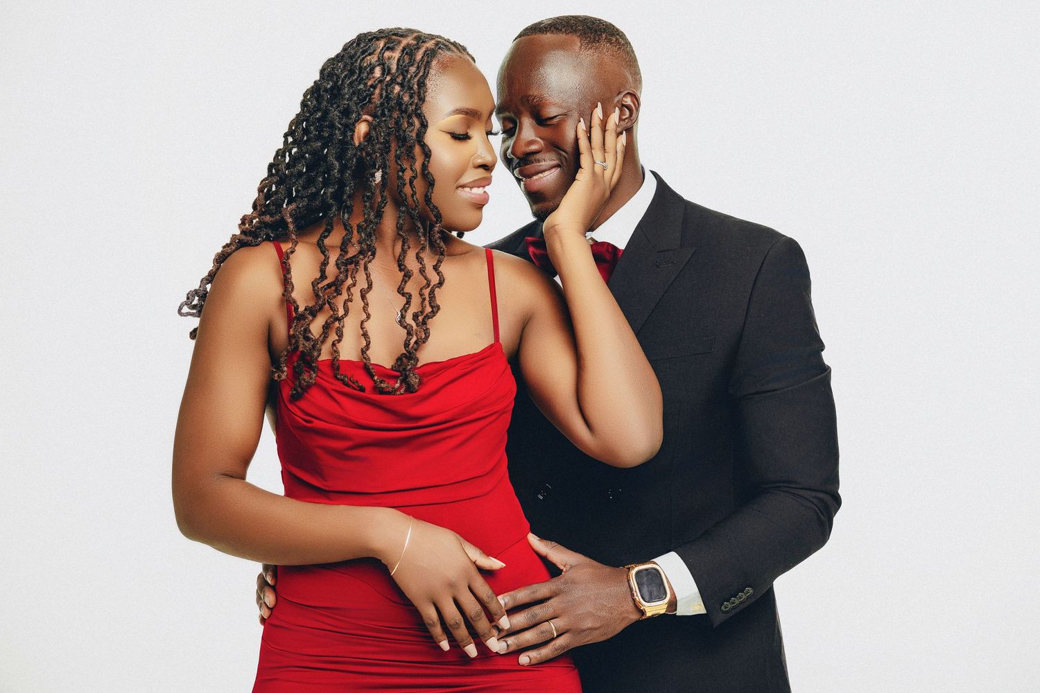 Romantic couple in matching red and black formal attire sharing an intimate moment against a white background.