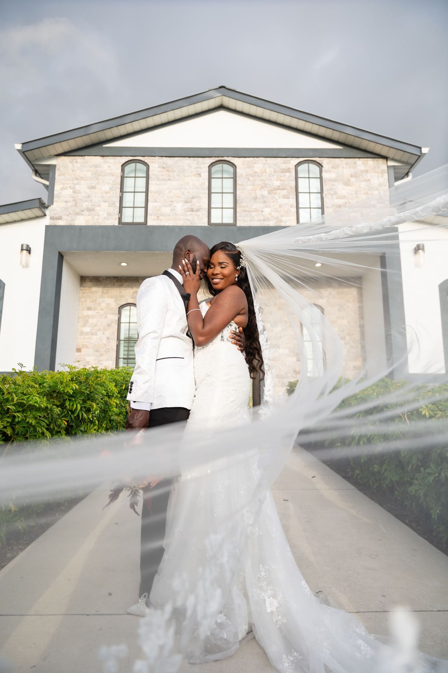 A bride and groom share a romantic moment outside a stone church, her veil flowing in the foreground.