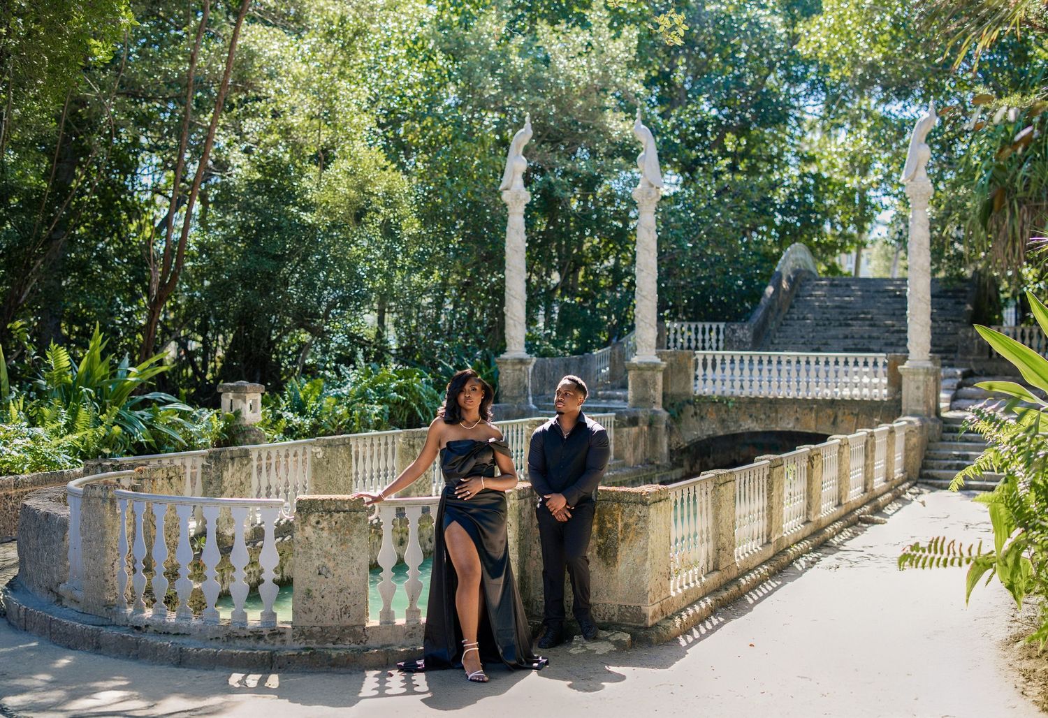 Couple in black formal wear posing at ornate stone balustrade in lush garden.