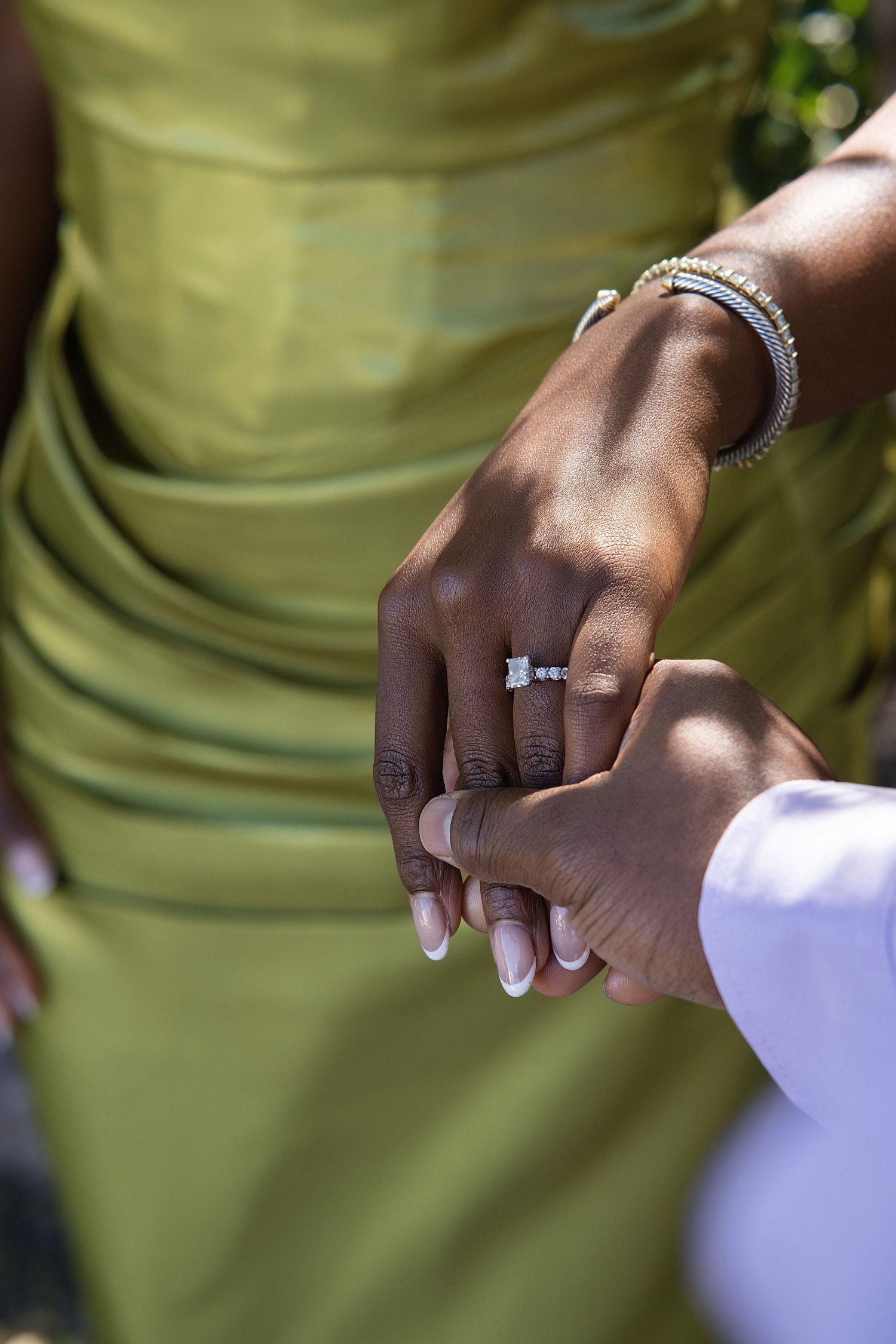 Close-up of Black woman\u2019s hand with diamond engagement ring held by partner, wearing green draped dress.