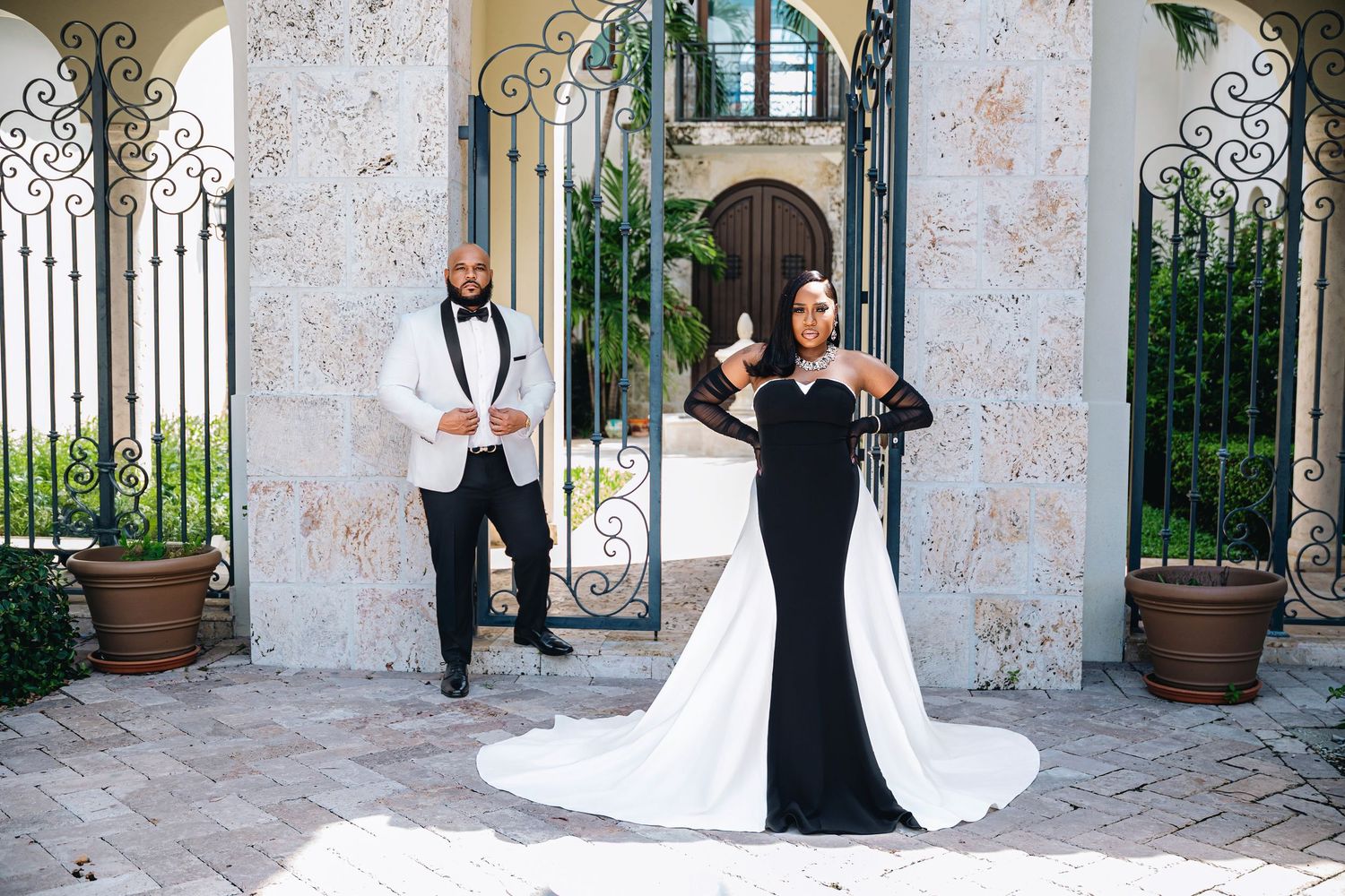 Elegant couple posing outside ornate gates, groom in white tuxedo, bride in stunning black and white gown with train.