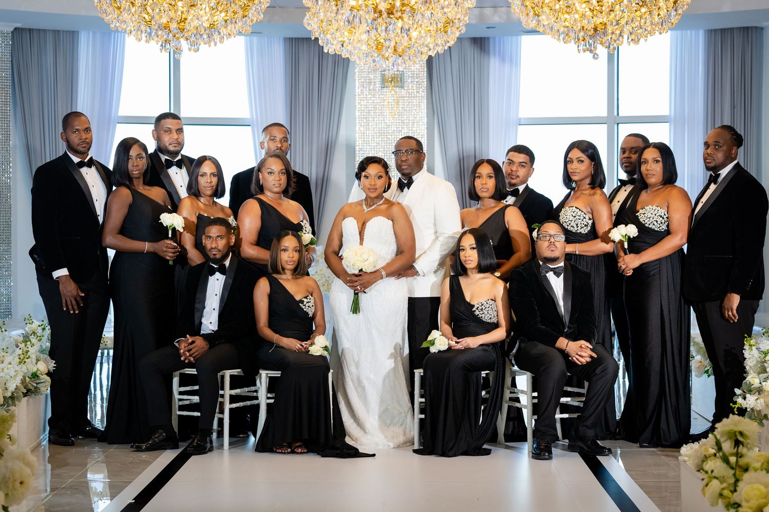 Wedding party in black attire poses with bride in white gown under chandeliers in elegant venue.