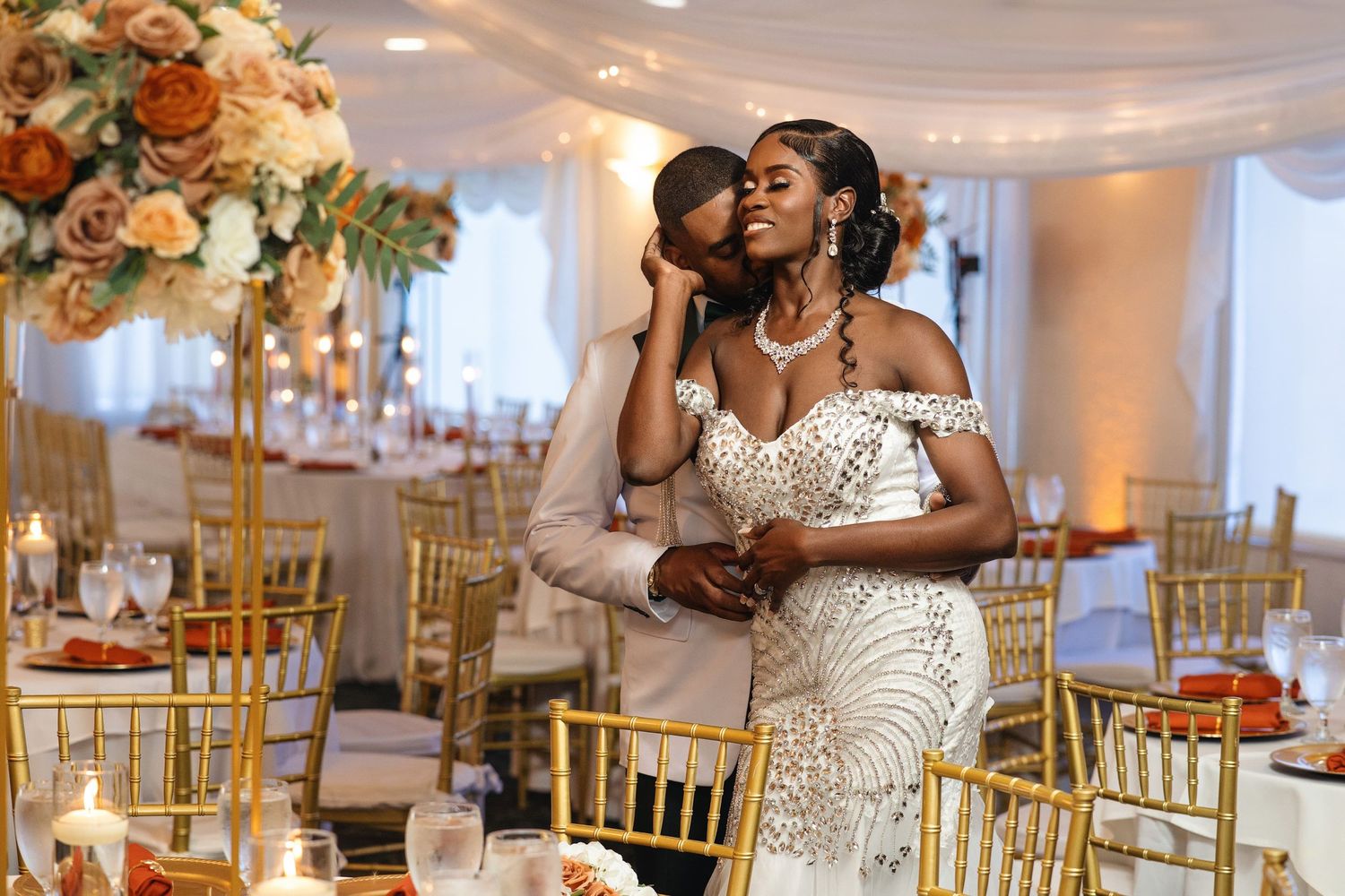 Elegant wedding couple embracing in a decorated reception hall with gold chairs and floral centerpieces.