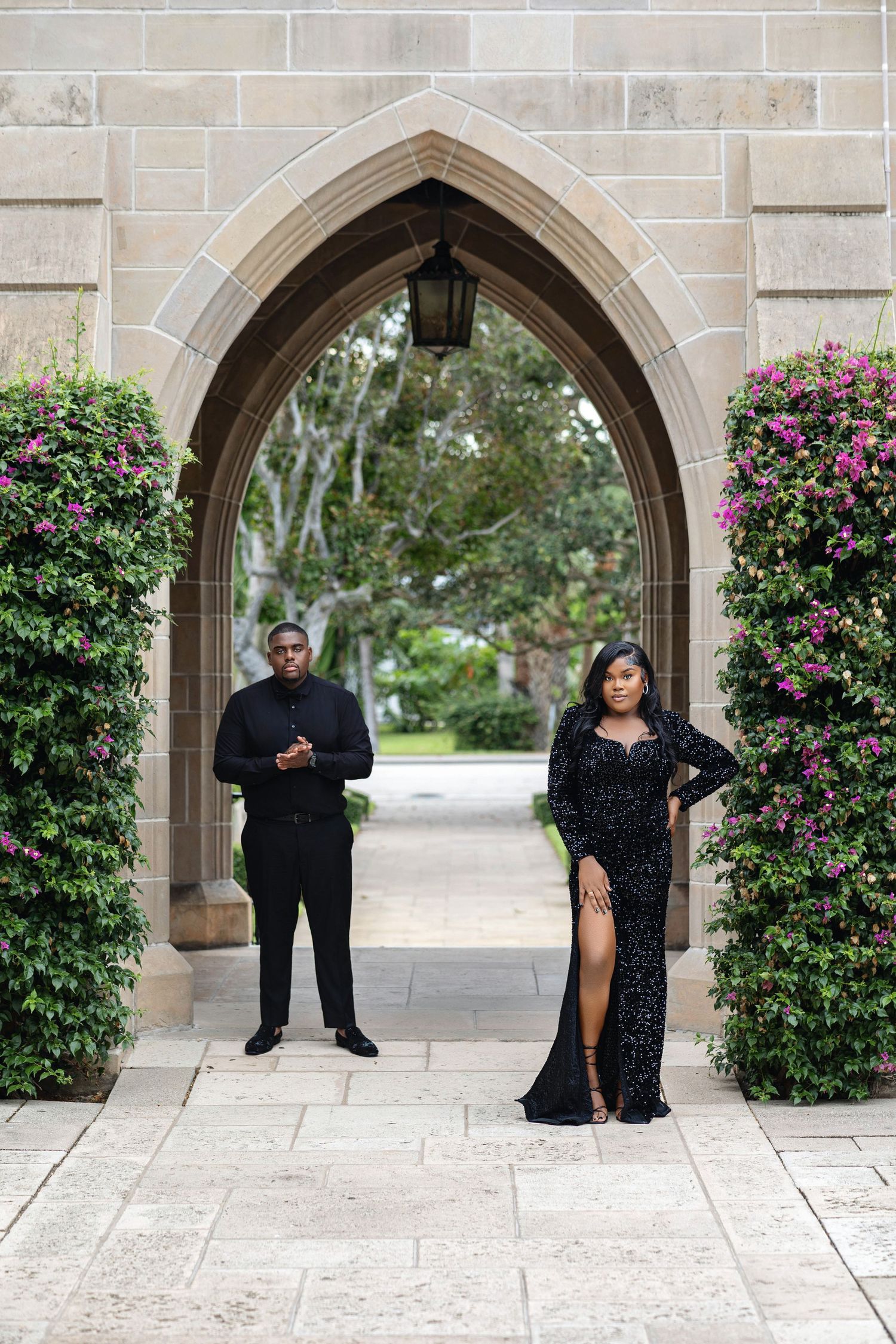 Couple in black formal wear standing under a Gothic stone archway adorned with flowering pink and purple vines.