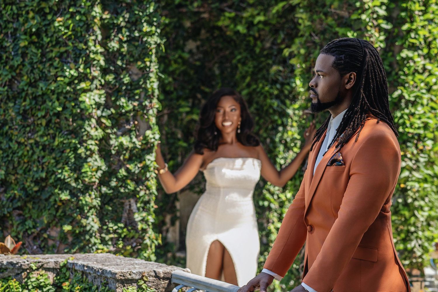 Bride-to-be in white dress reaching for groom in orange suit near ivy wall at outdoor venue.