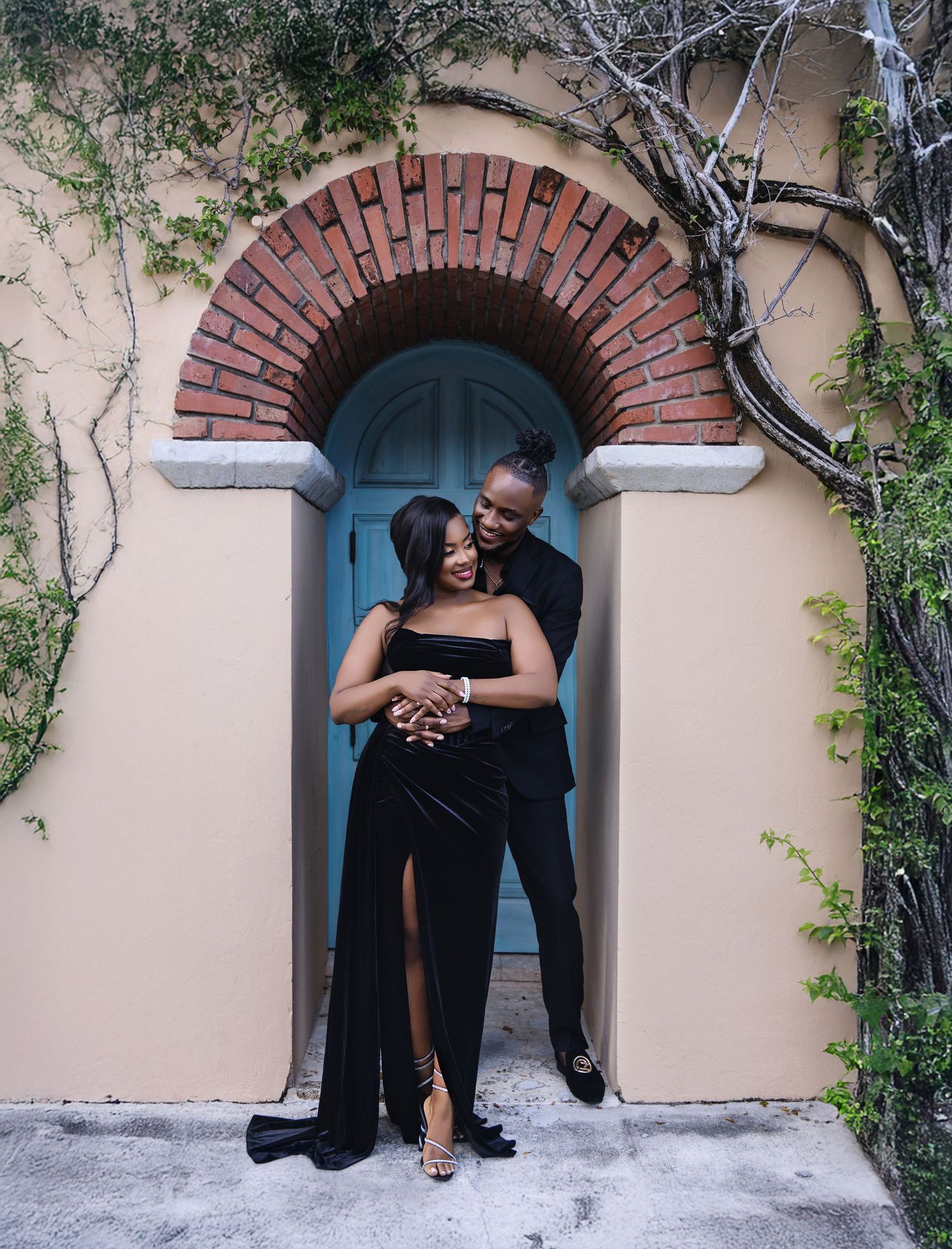Couple in black formal attire embracing under a brick arch doorway outdoors.