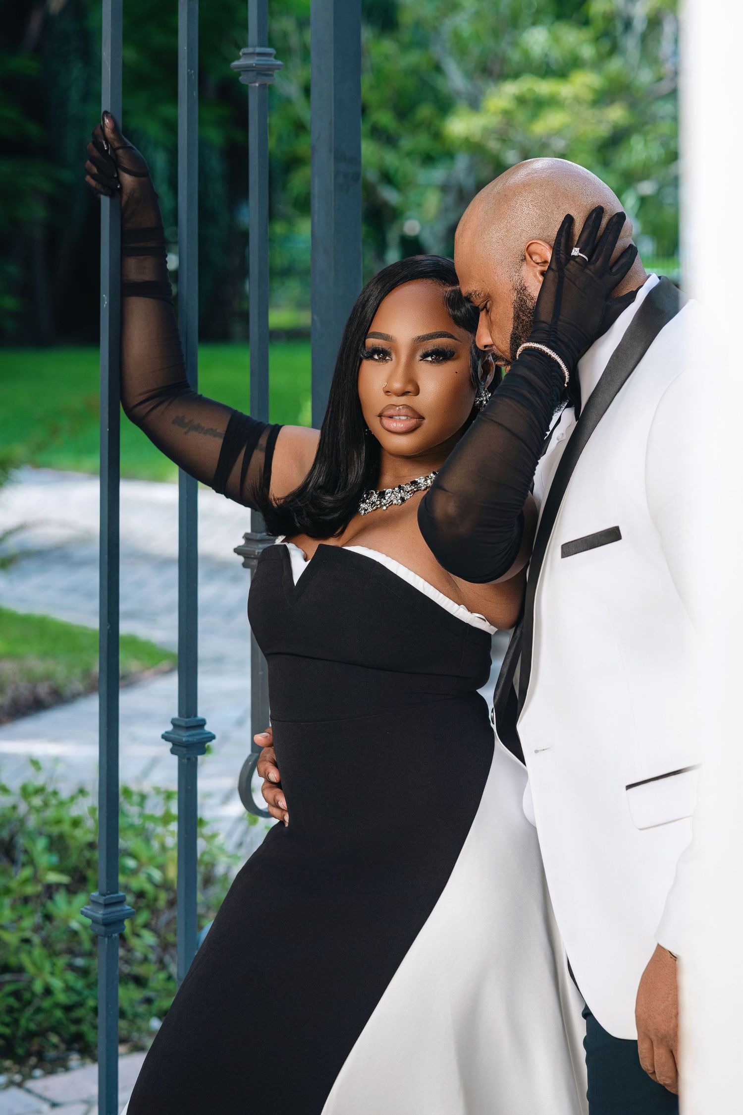 Black couple in black and white formal wear lean against ornate iron gate in garden setting.