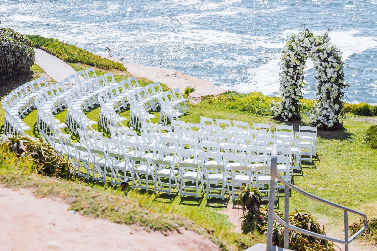 White chairs arranged in curved rows for an outdoor wedding ceremony by the ocean, with a floral arch near the cliff edge.