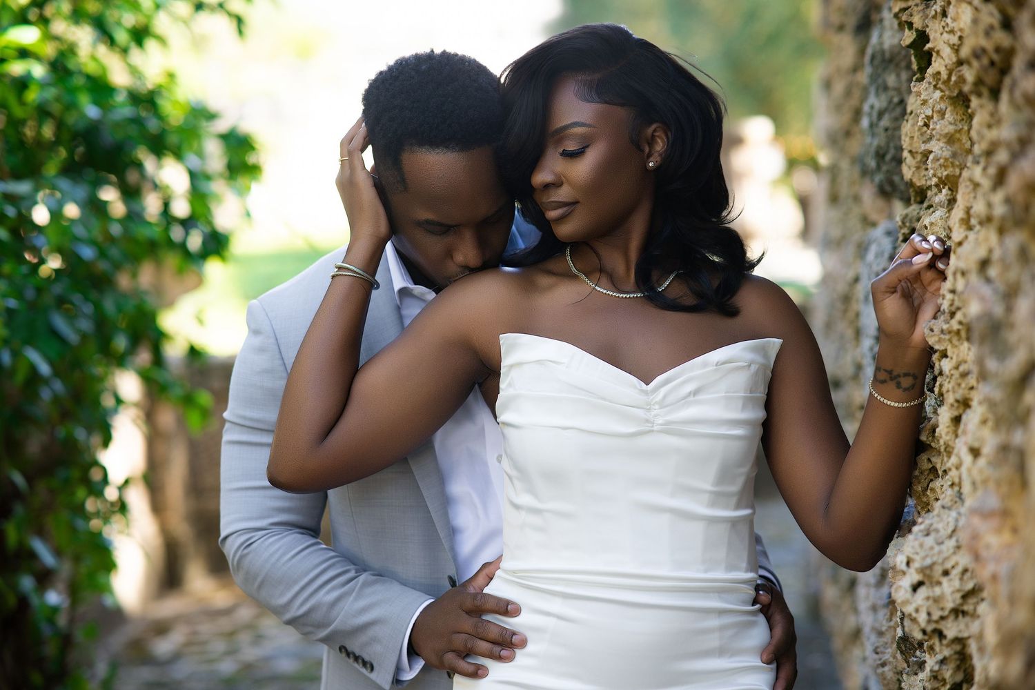 Bride in white strapless gown and groom in gray suit in intimate embrace by stone wall.