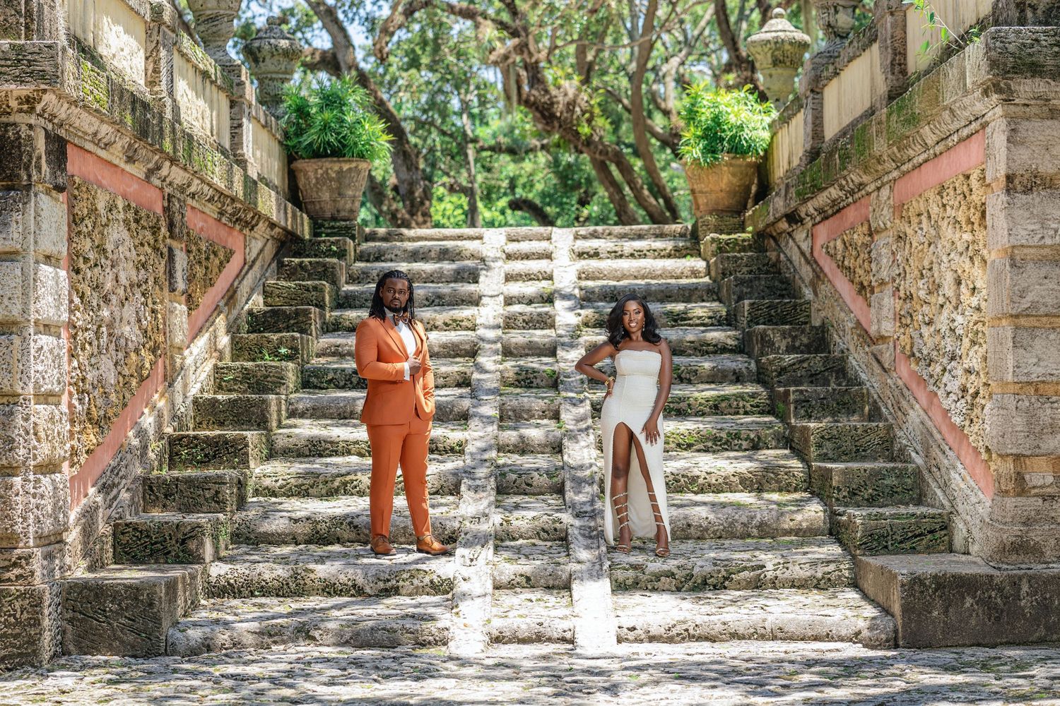 Two women in orange and white outfits posing on ornate moss-covered stone staircase.