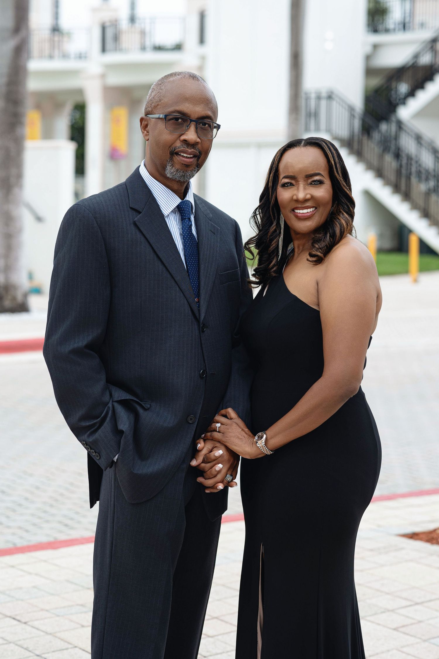 Stylish engaged couple holding hands and posing together outdoors, dressed in black evening wear and navy suit.