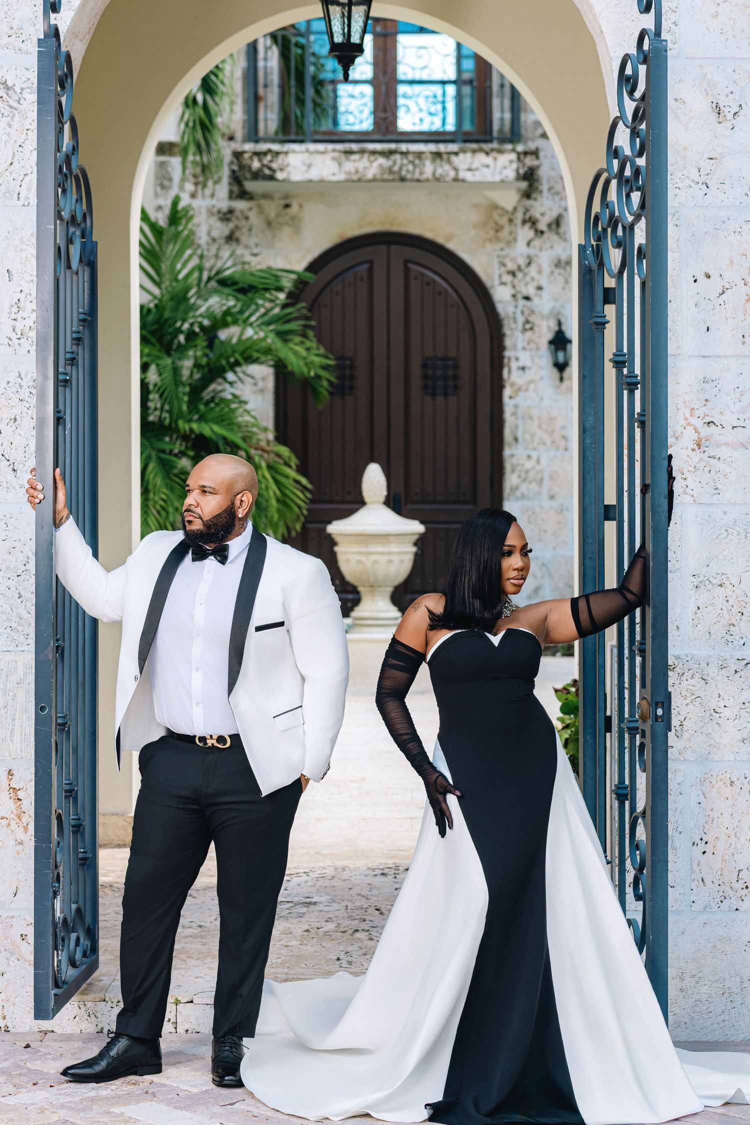 Stylish couple poses at ornate blue iron gates of a luxury estate, wearing black and white formal attire.