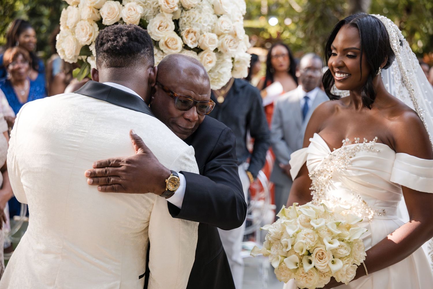 Groom in white suit hugs guest at outdoor wedding while bride in off-shoulder gown holds white bouquet.