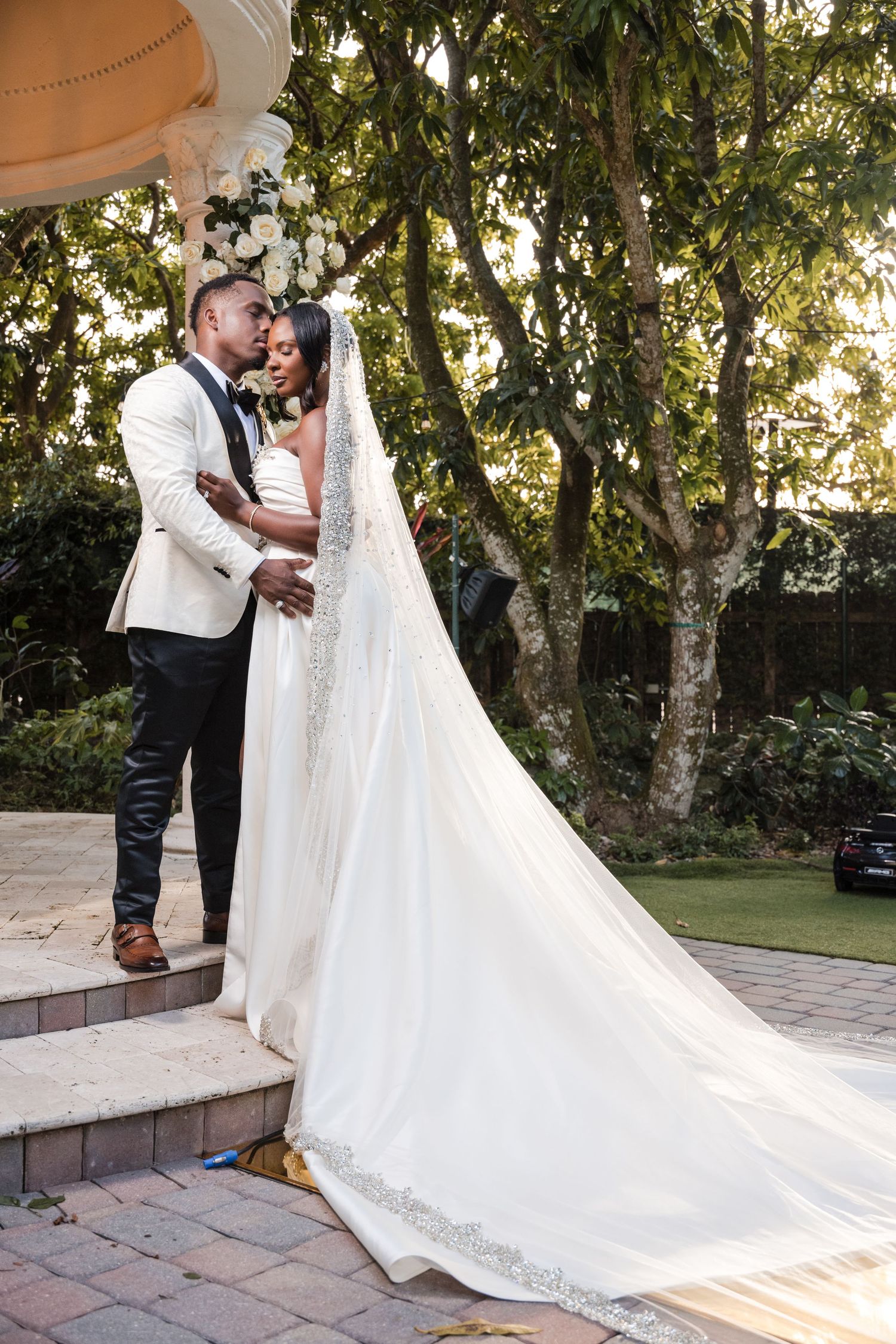 Bride and groom share a romantic moment on steps, bride wearing a long white veil and gown at outdoor wedding ceremony.