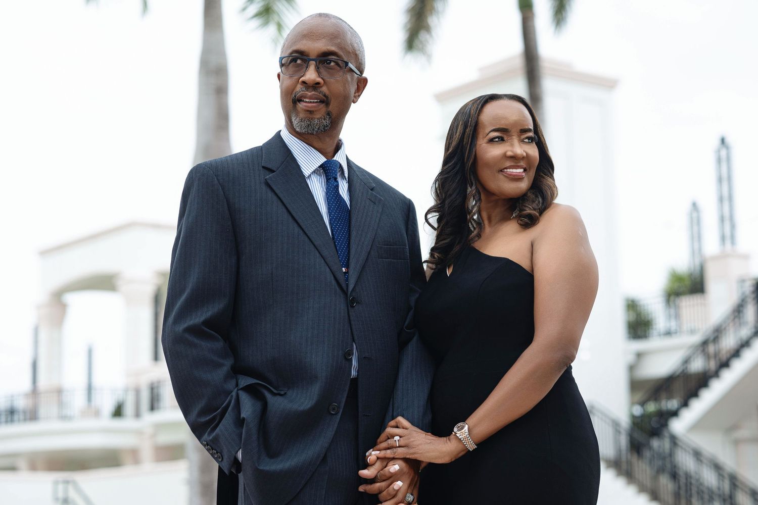 Engaged couple holding hands and posing together near white columns, dressed in formal black and navy attire.
