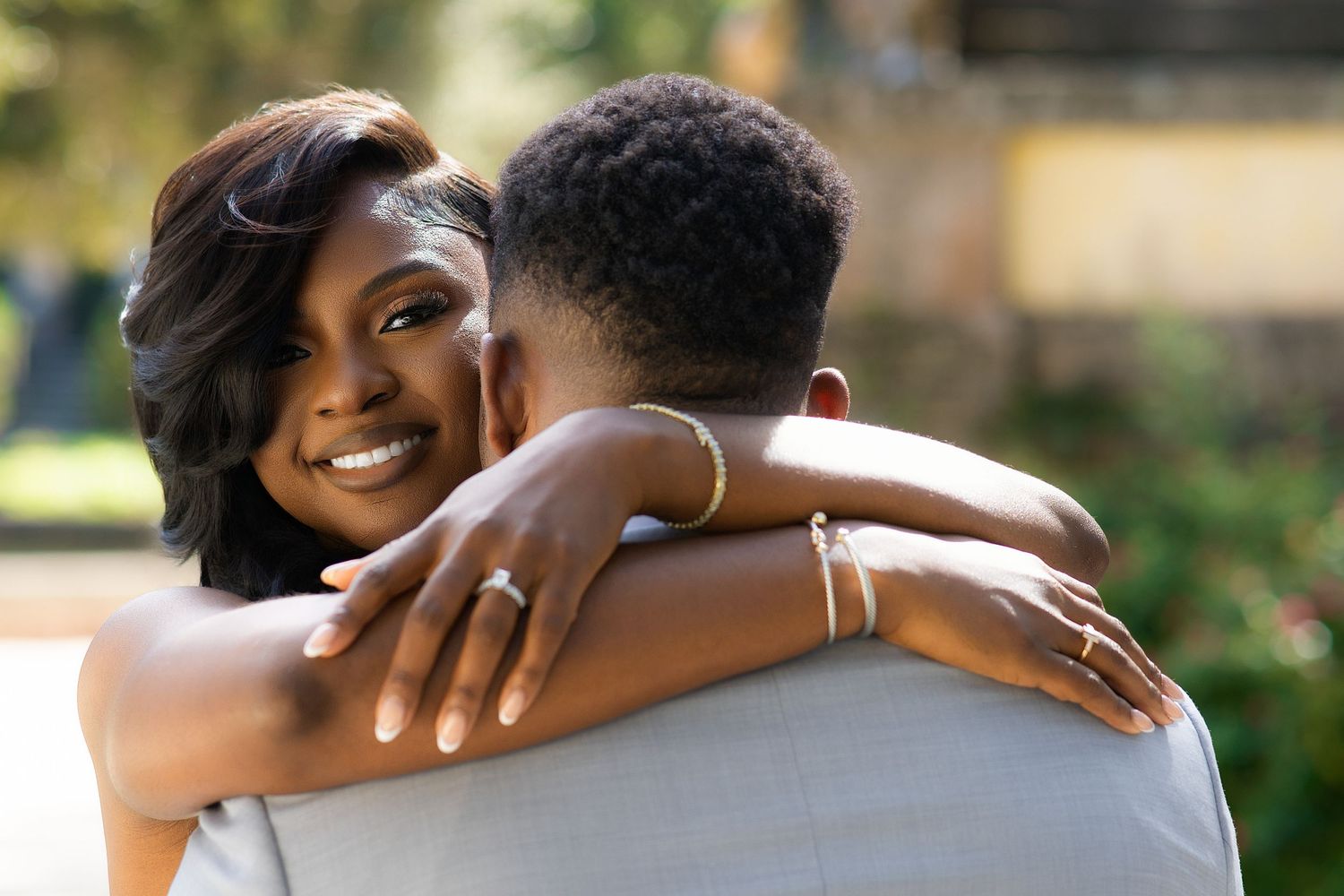 Joyful Black bride hugging groom while showing off diamond engagement ring, smiling outdoors.