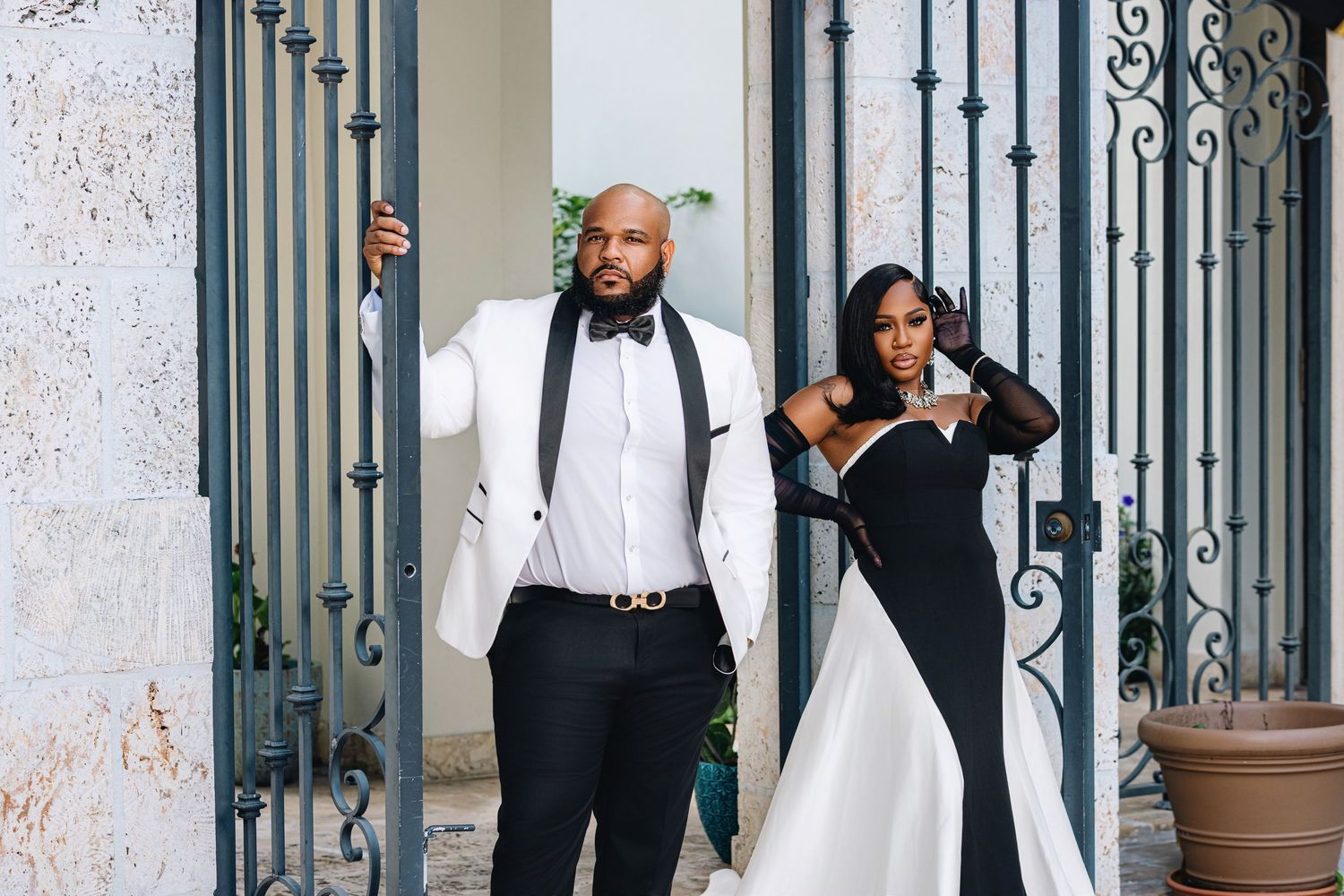 Stylish wedding couple posing at iron gates, groom in white blazer and black bow tie, bride in sleek black gown.